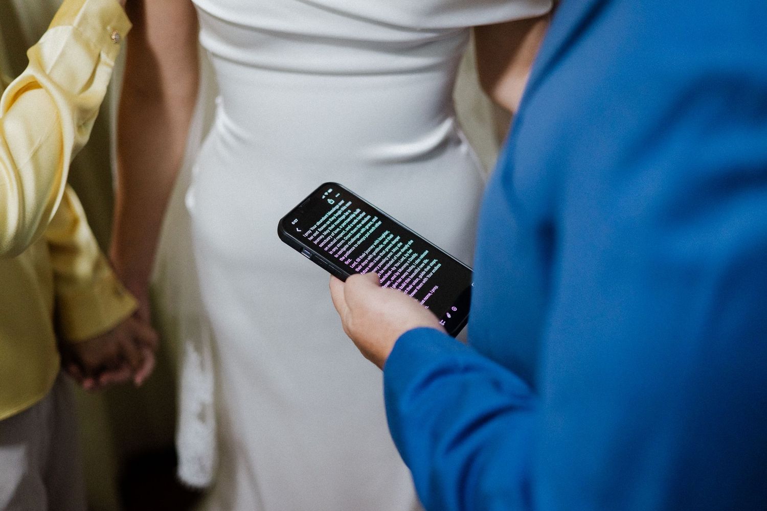 Person in blue jacket holds smartphone displaying colorful audio waveform near bride in white wedding dress.