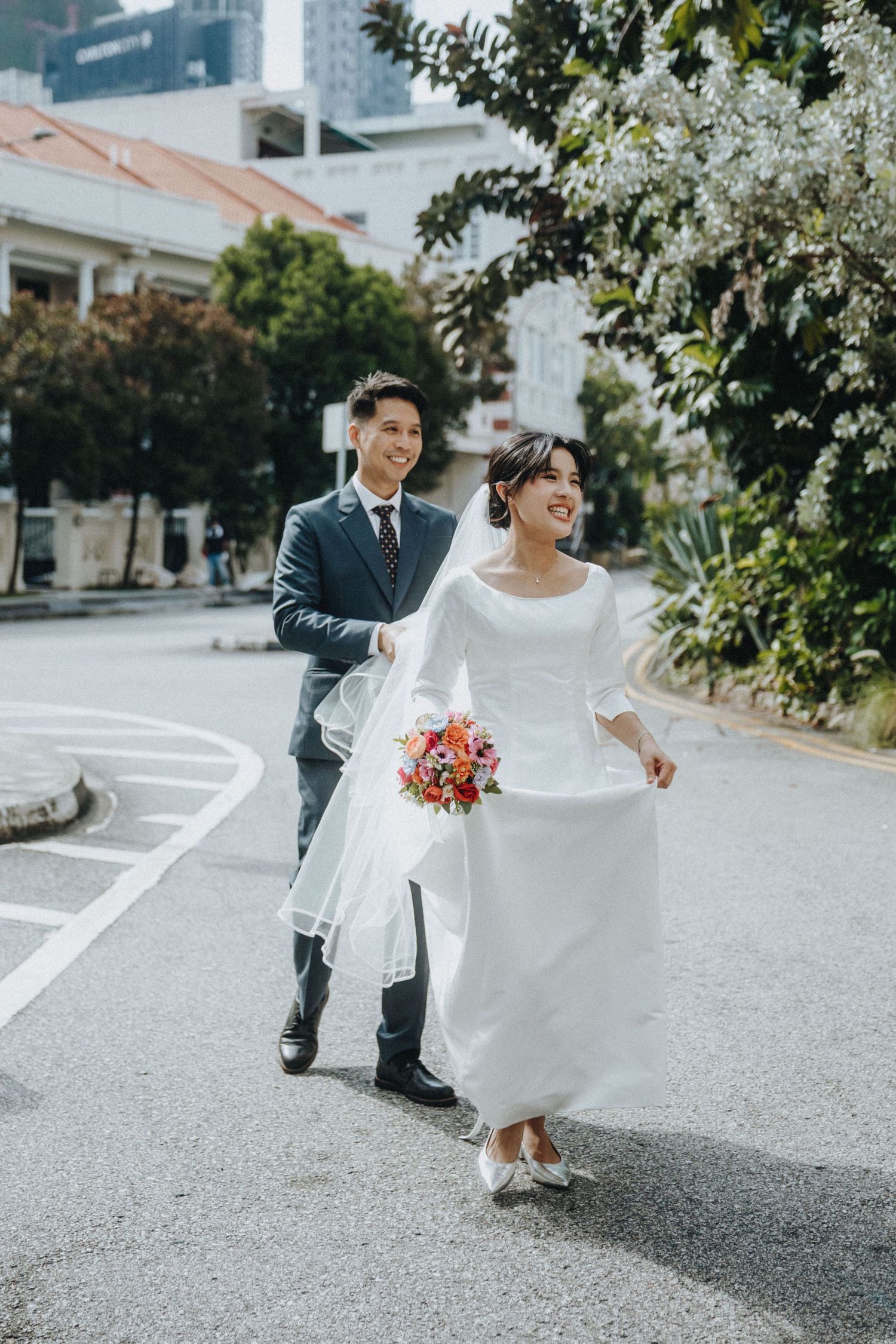 Smiling bride in white gown walks with groom holding her veil on a sunny Singapore street.