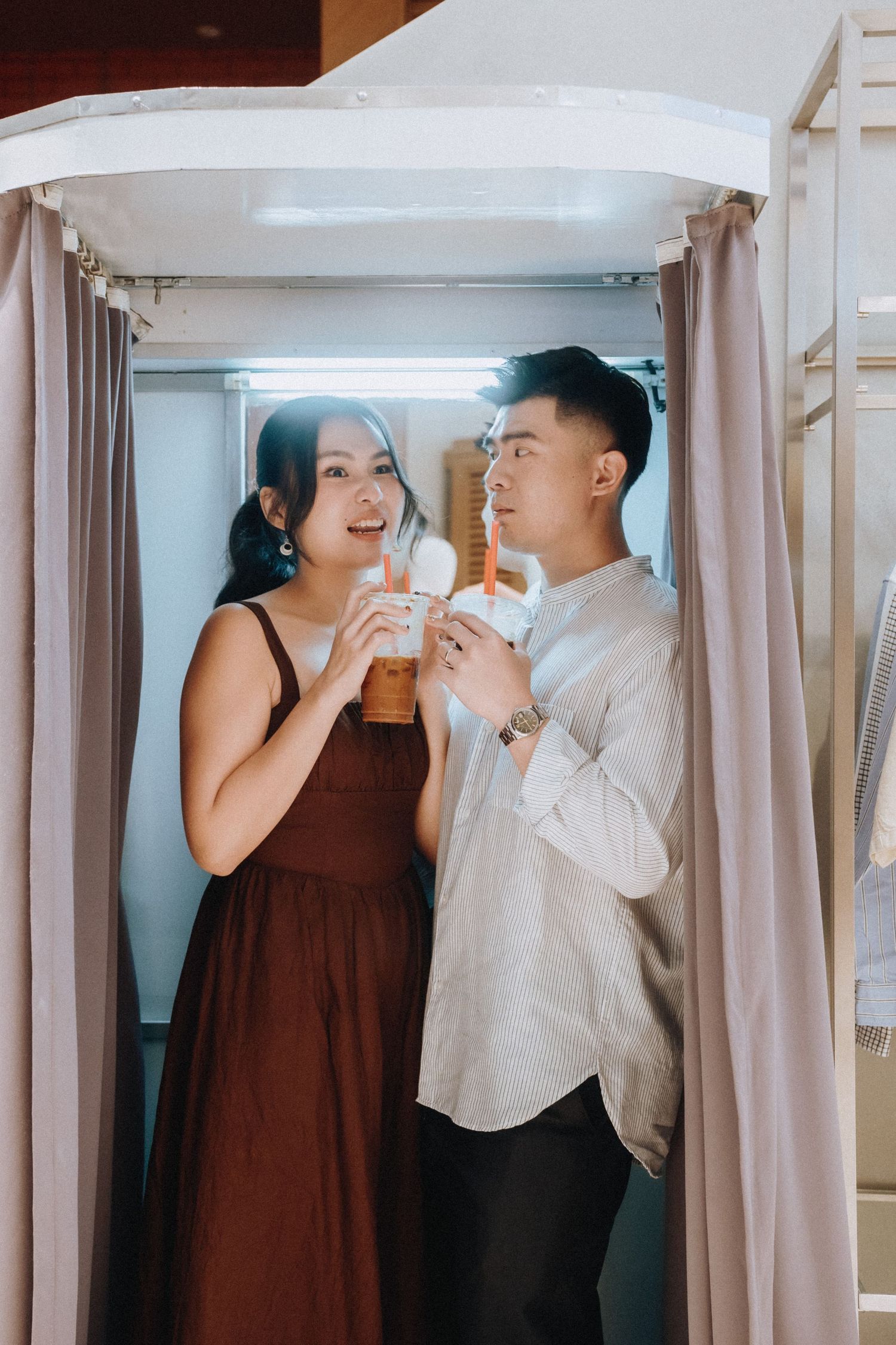 Happy Asian couple toasting with iced drinks in a photo booth with purple curtains.