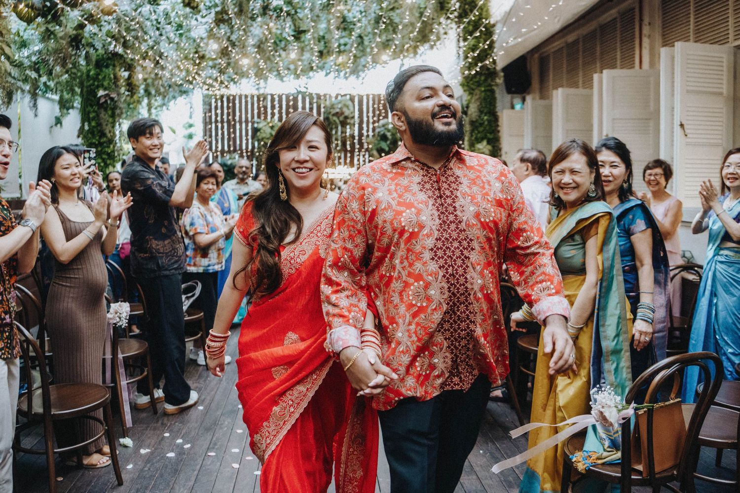 Newlyweds in matching red Indian attire walk joyfully down aisle at multicultural wedding.