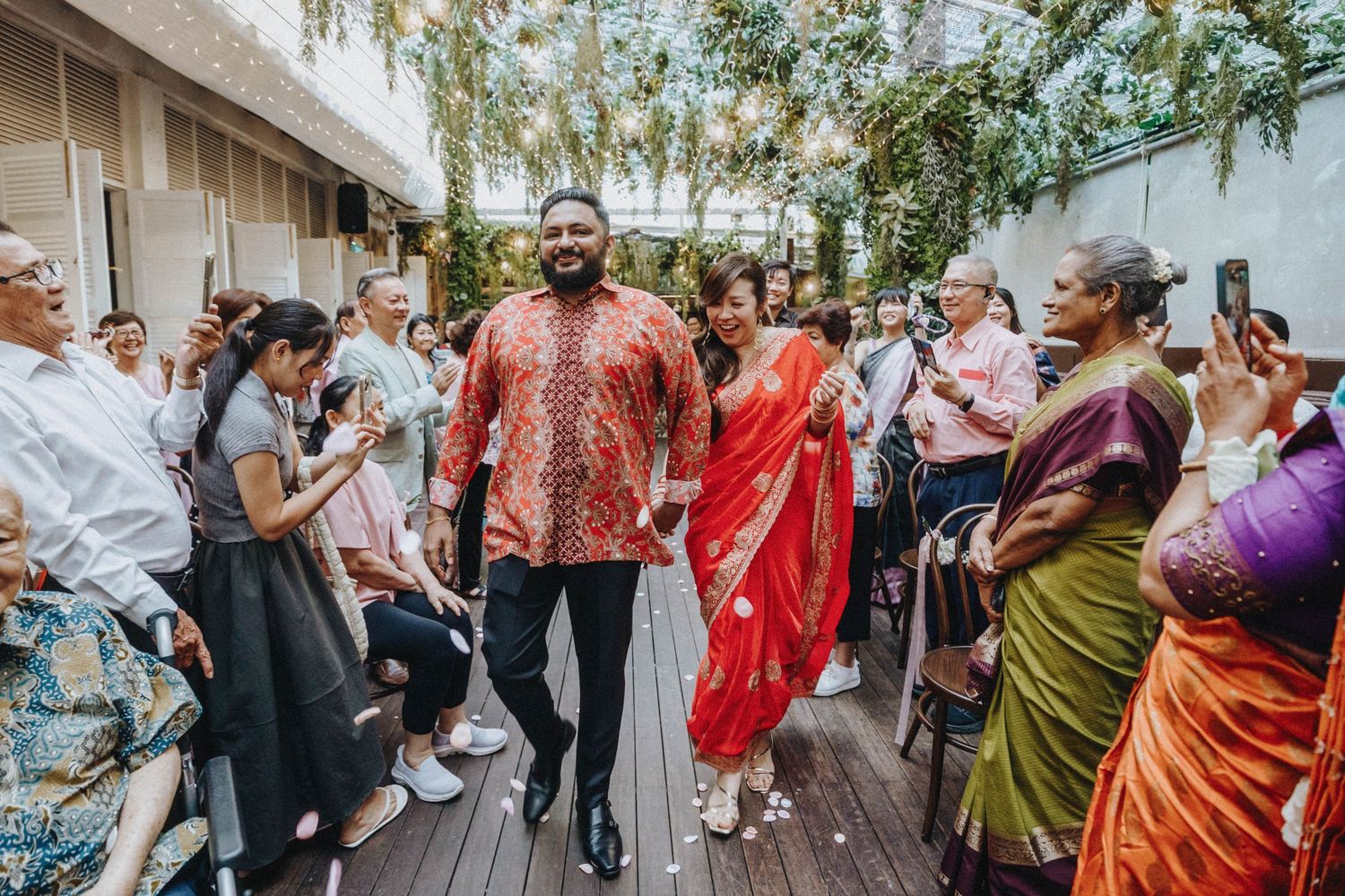 Couple in vibrant traditional attire walks through cheering guests at an outdoor wedding celebration.