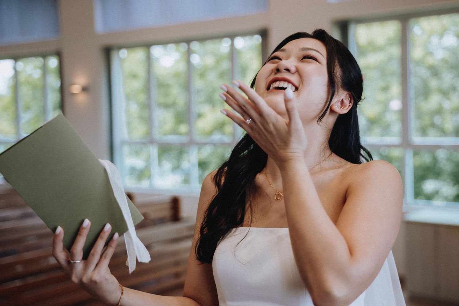 Joyful bride in white dress laughing while reading a heartfelt note, holding it up near bright windows.