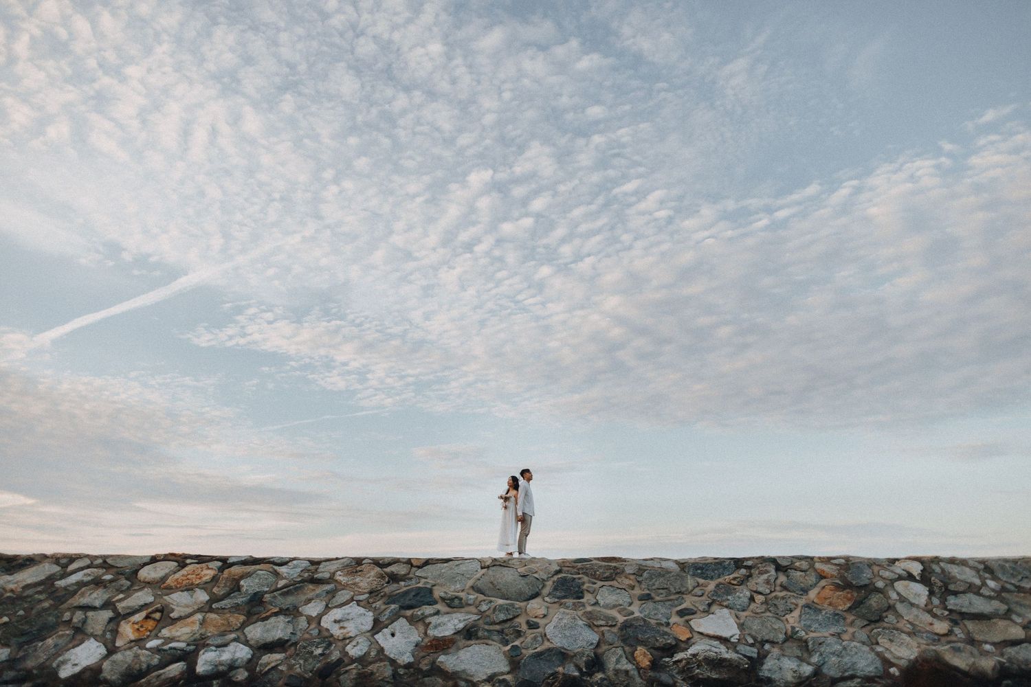 A couple stands on rocky terrain against a dramatic cloudy sky during sunset.