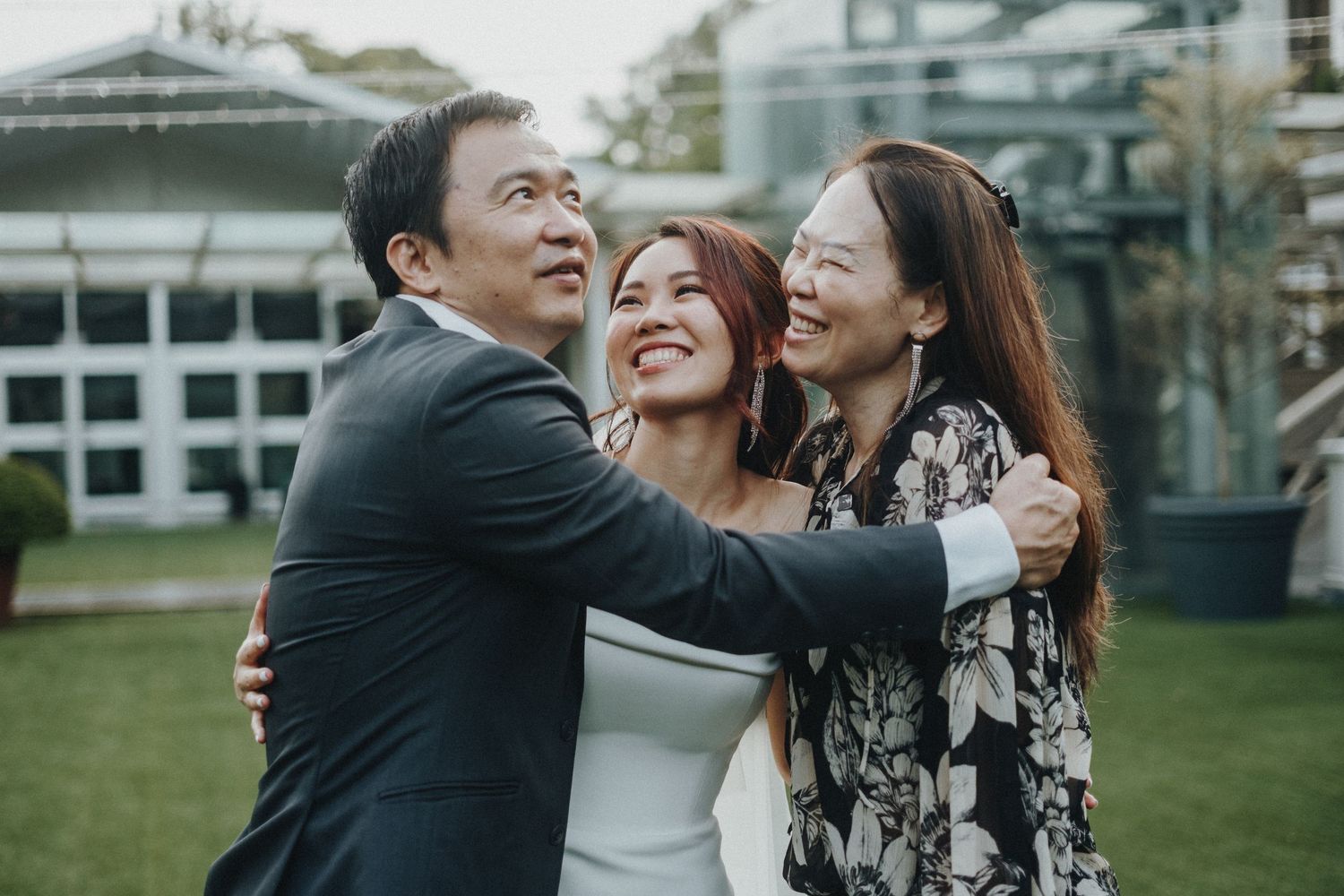 Three people embrace and laugh joyfully together on a green lawn with buildings in the background.