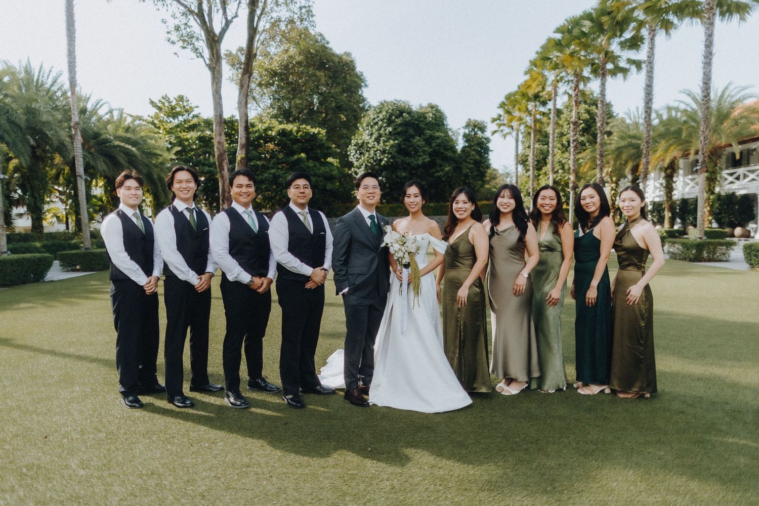Wedding party poses on a green lawn with palm trees, groomsmen in black vests and bridesmaids in neutral gowns.