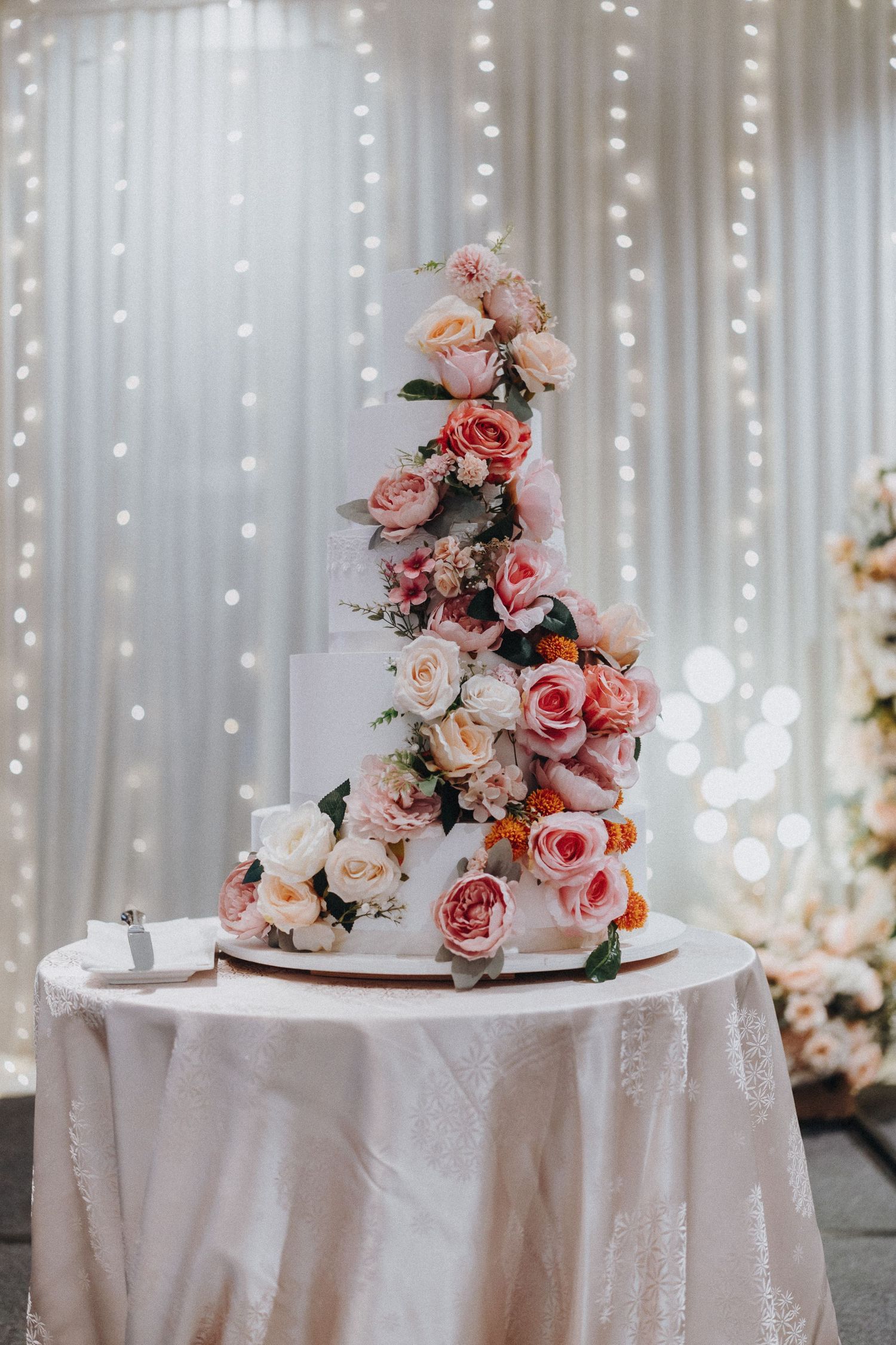Elegant wedding cake decorated with cascading pink and peach roses against a backdrop of twinkling lights.
