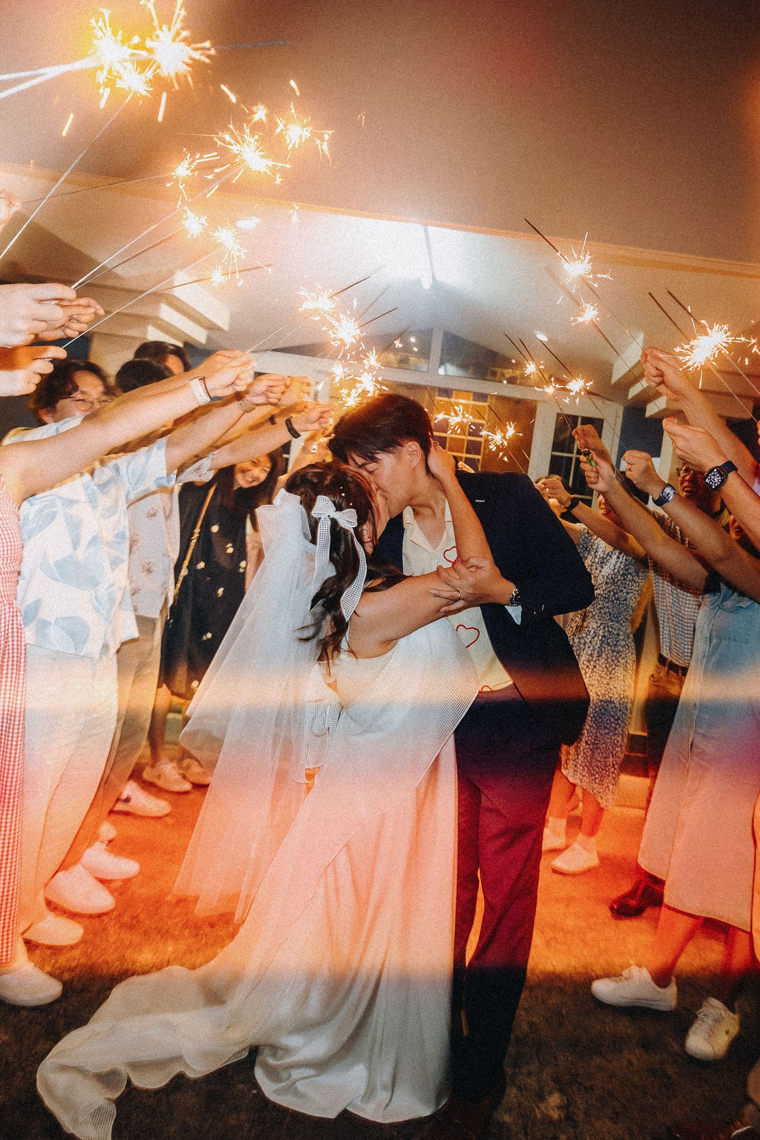 Wedding guests holding sparklers creating light streaks in dimly lit venue.