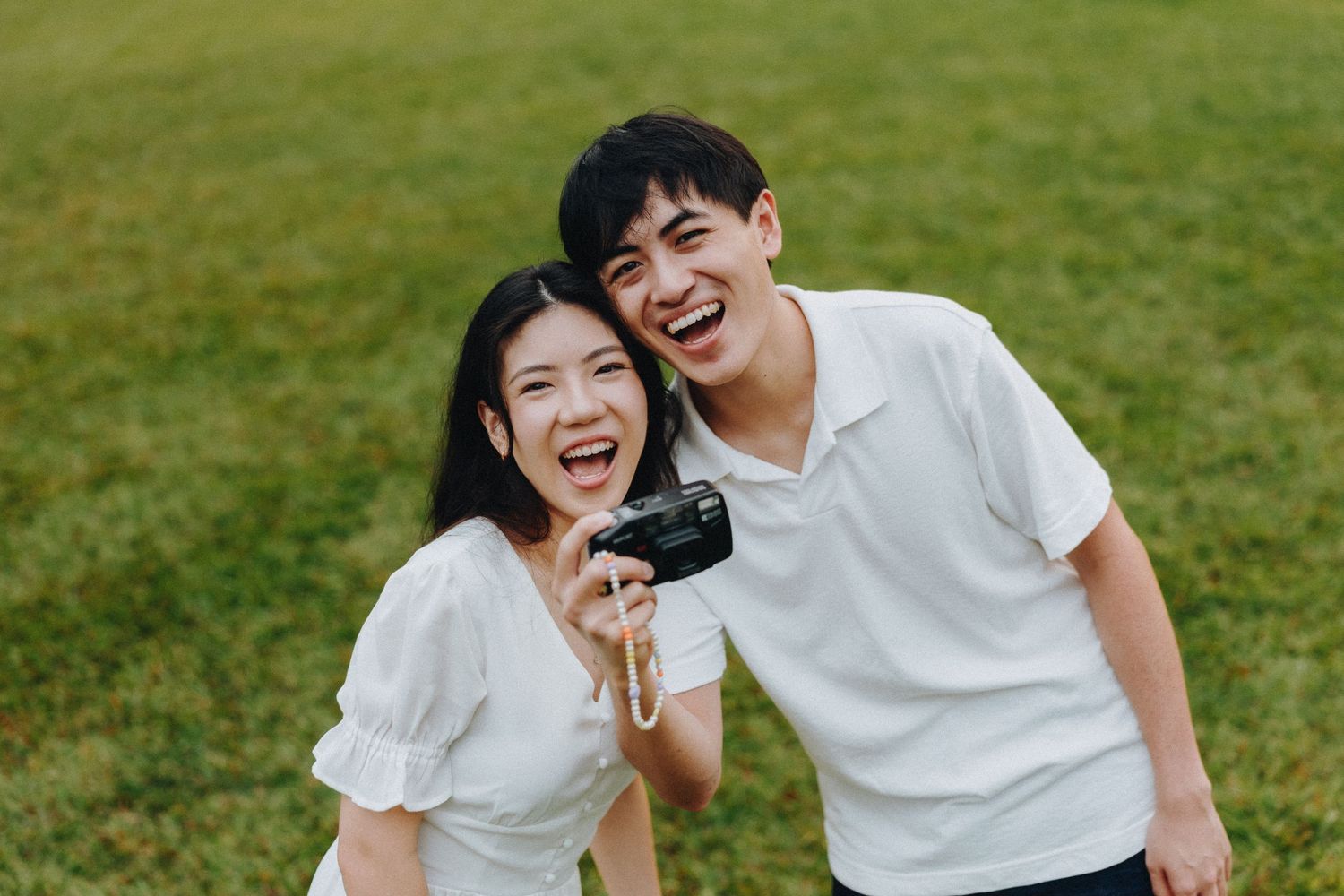 Happy young couple shares joyful moment outdoors while holding a camera on grassy lawn.