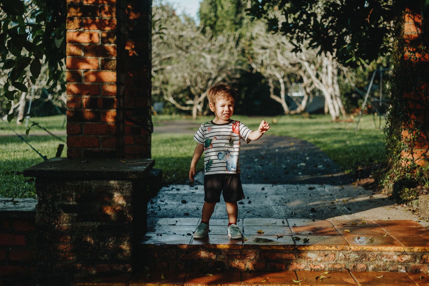 A toddler in stripes stands on a path surrounded by dappled sunlight and trees.