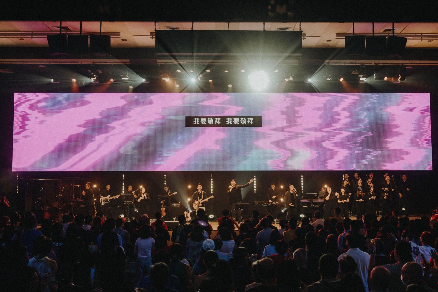 A wide concert venue showing a large display screen with pink graphics above an orchestra section.