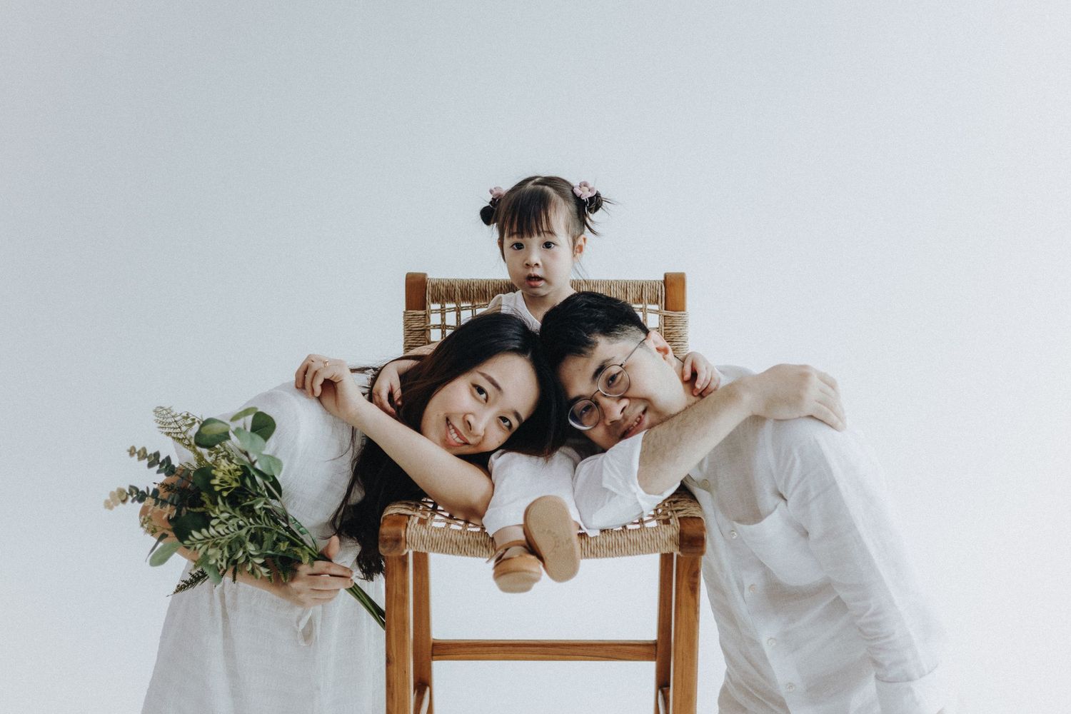 Playful family photo with wooden chair and greenery against white backdrop.