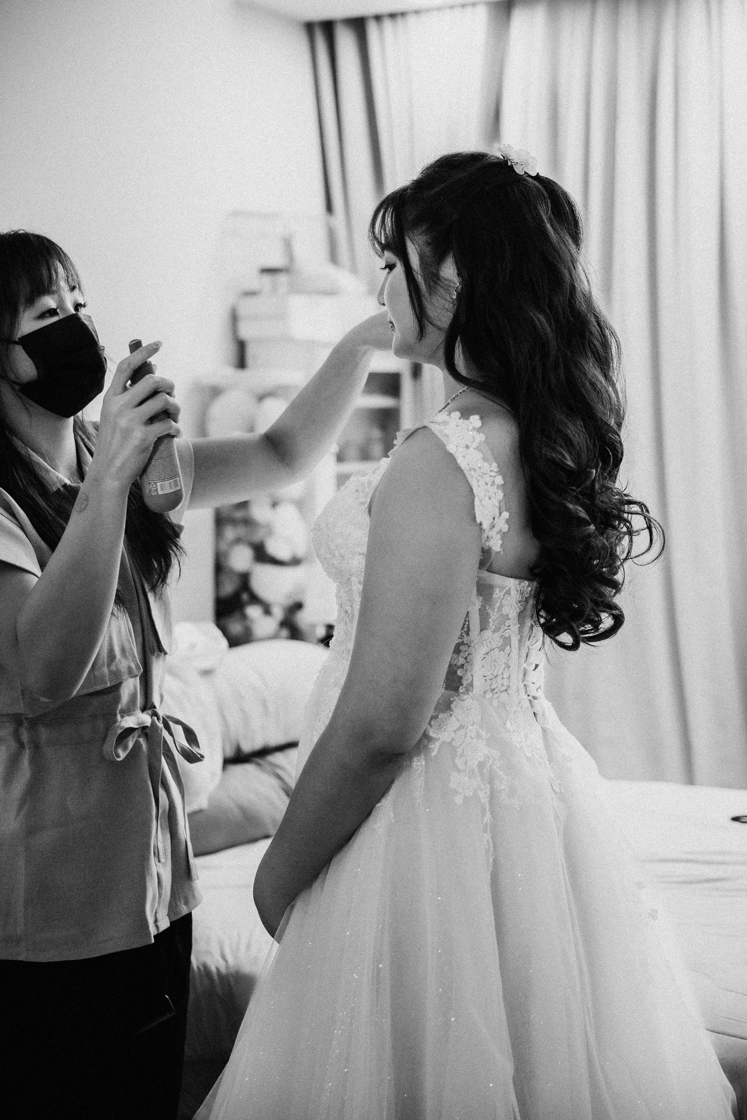 A stylist in a black mask arranges a bride's elegant white lace wedding dress during preparation.