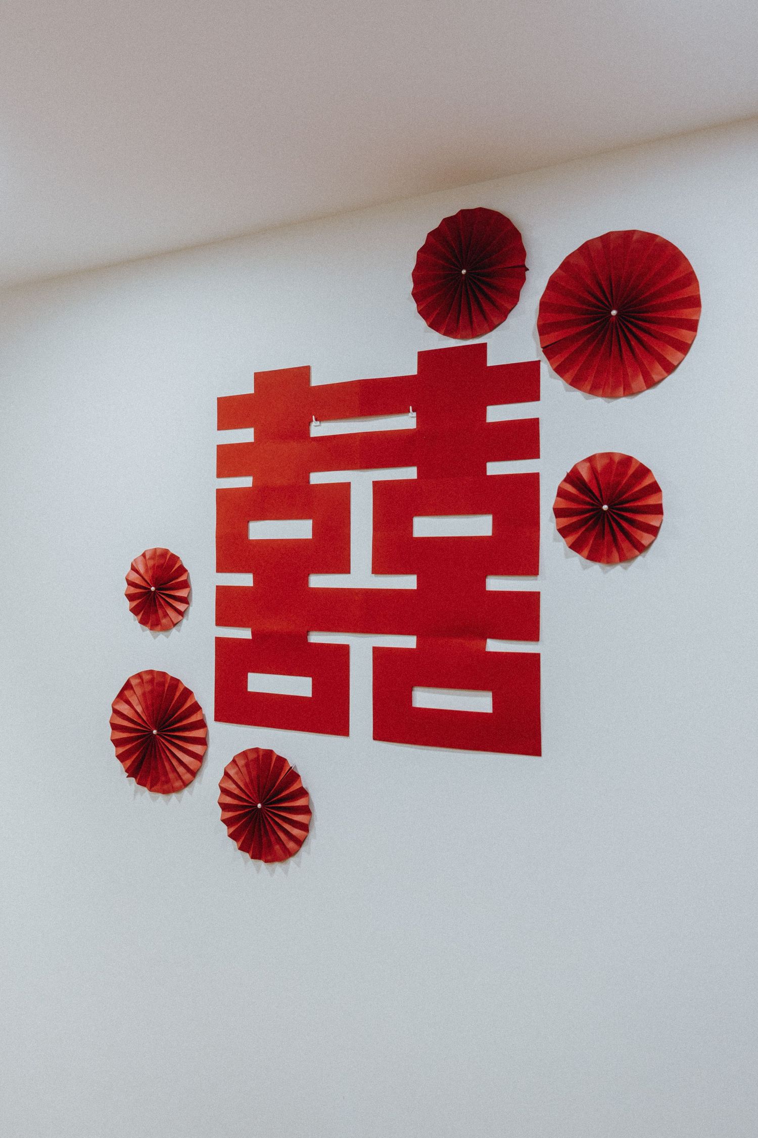 Red double happiness symbol with red paper fan decorations on white wall for traditional Chinese wedding decor.