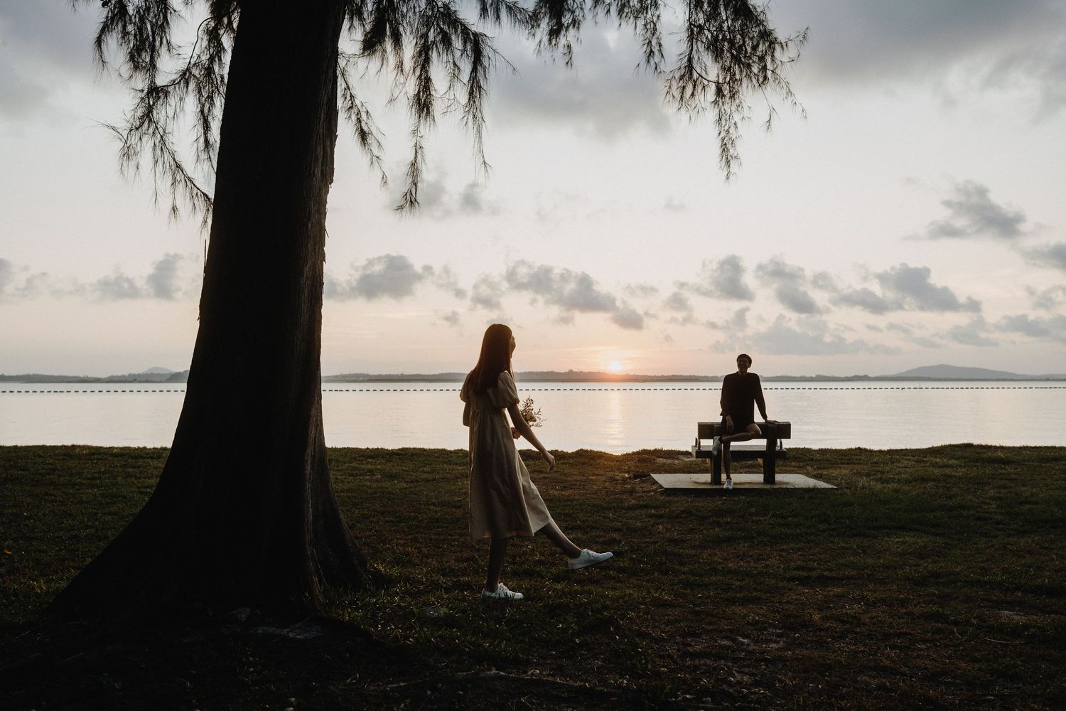 Silhouette of a person walking dog at sunset while another person sits on bench by lake under a large tree.