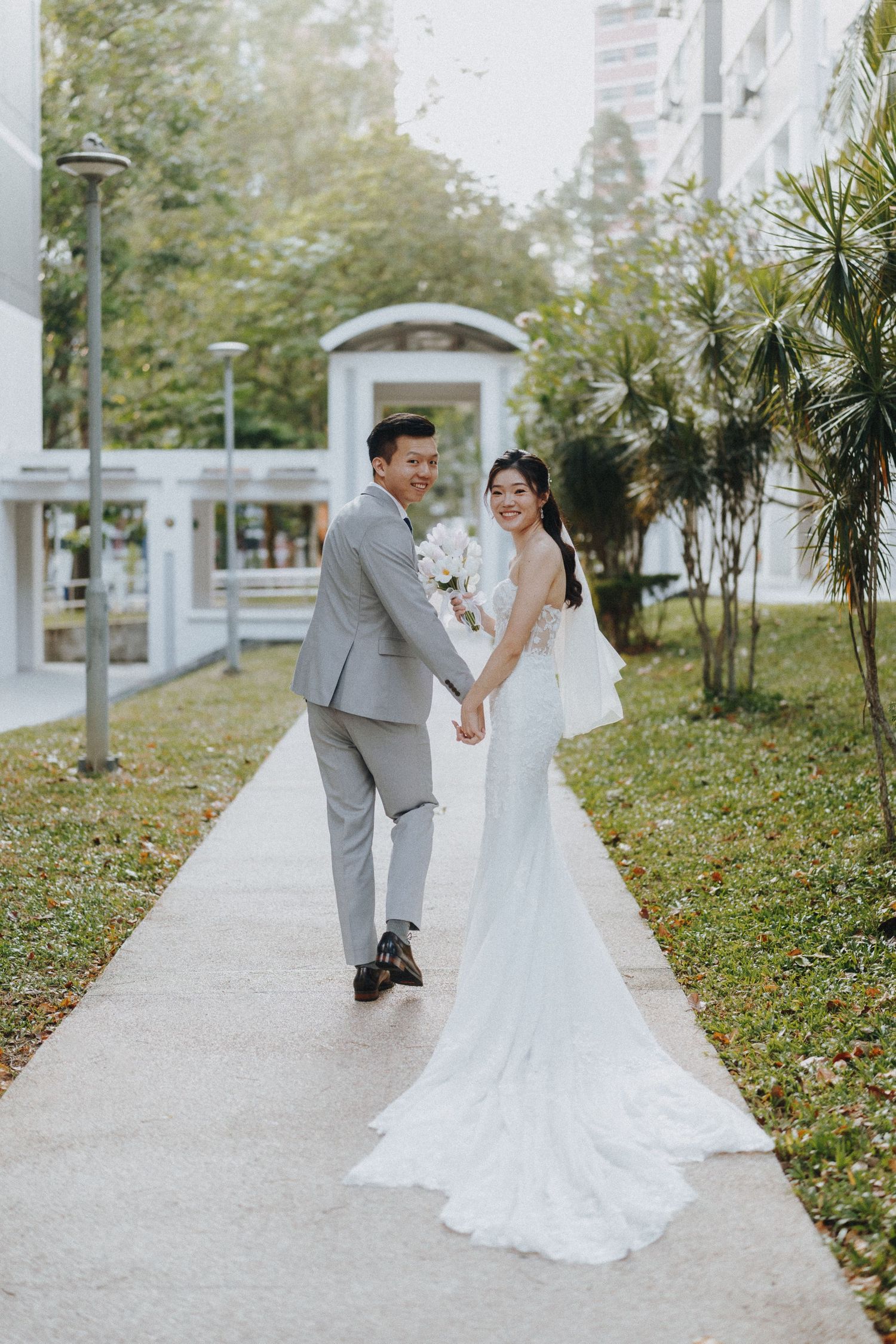 Couple in wedding attire walking and posing on an outdoor pathway surrounded by greenery.