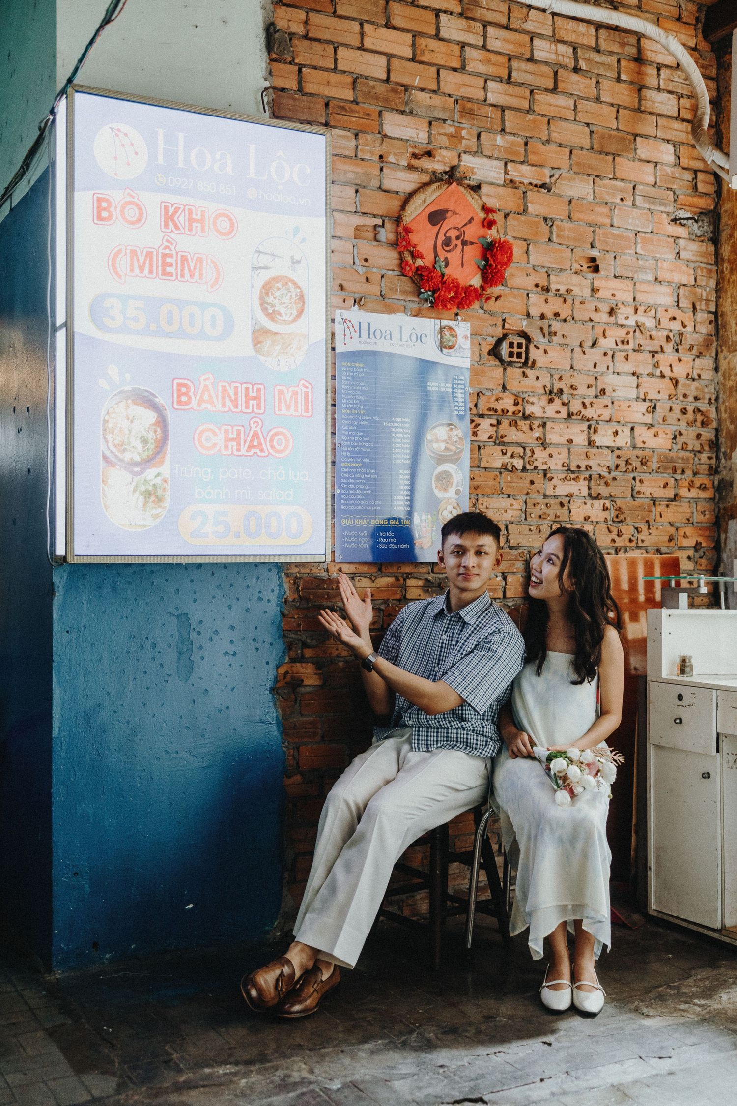 A couple sits outside a Vietnamese restaurant with a menu board displaying Banh Mi Chao on a brick wall.