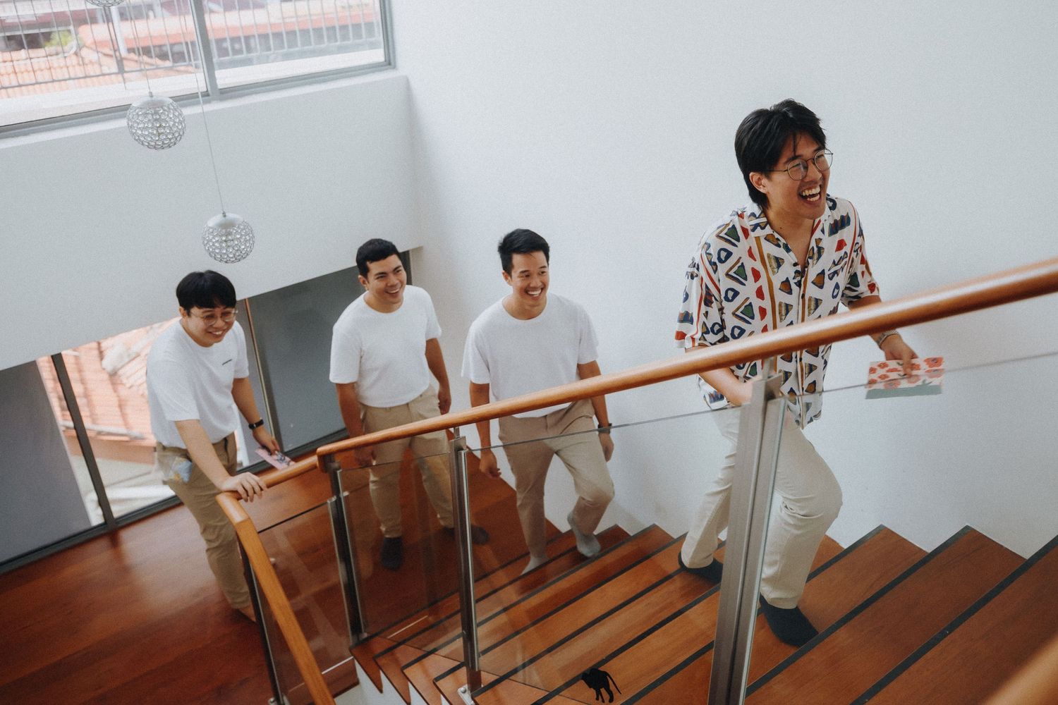 People in white outfits and a person in printed shirt walking up wooden stairs with handrails in a bright interior space.