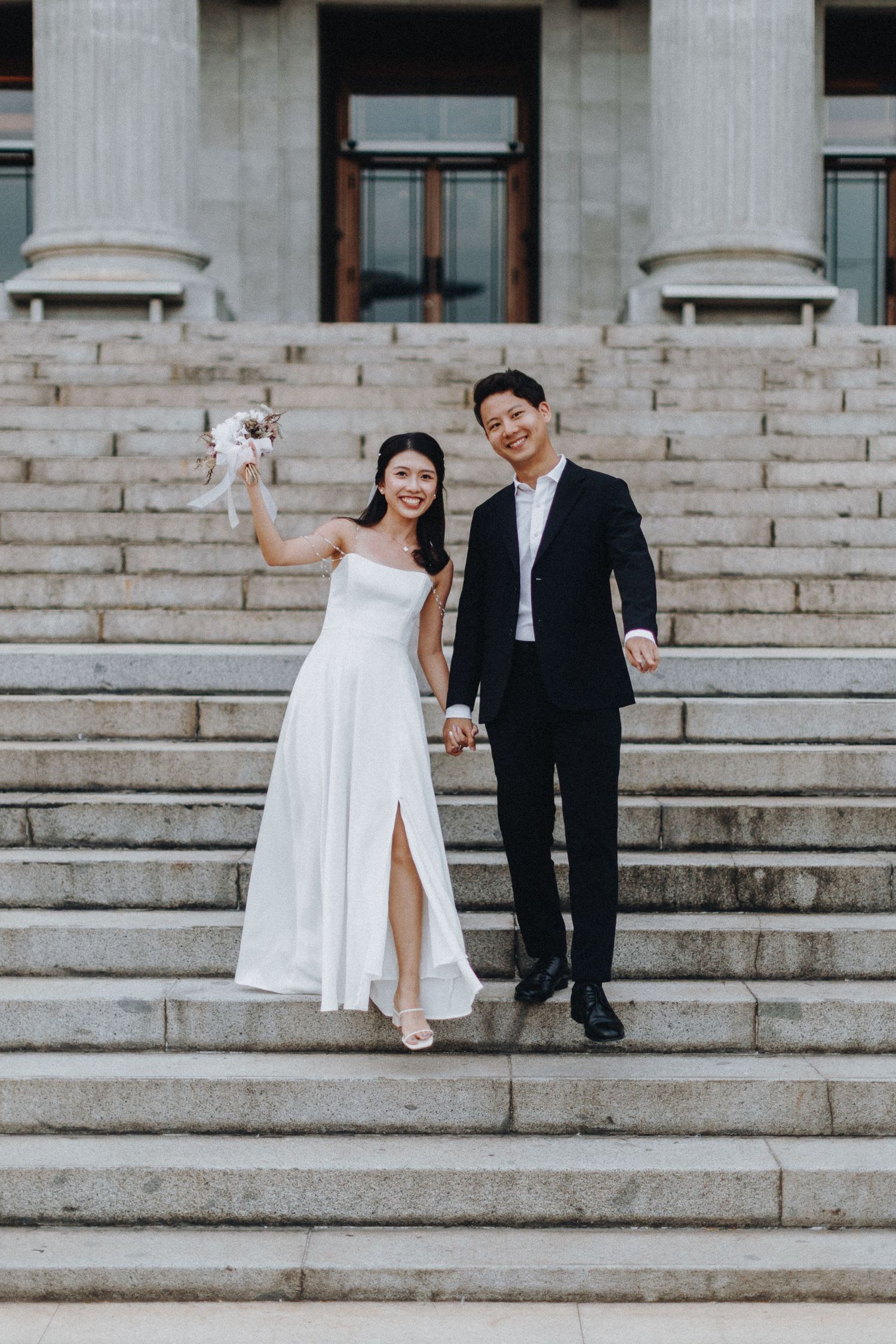 A couple celebrates on grand stone steps in formal wedding attire.