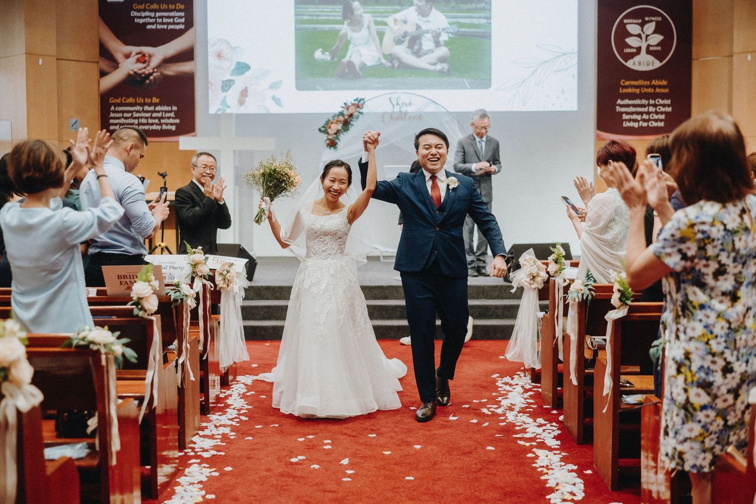 Joyful newlyweds walk down church aisle lined with red carpet and pews as guests celebrate with white flower petals.