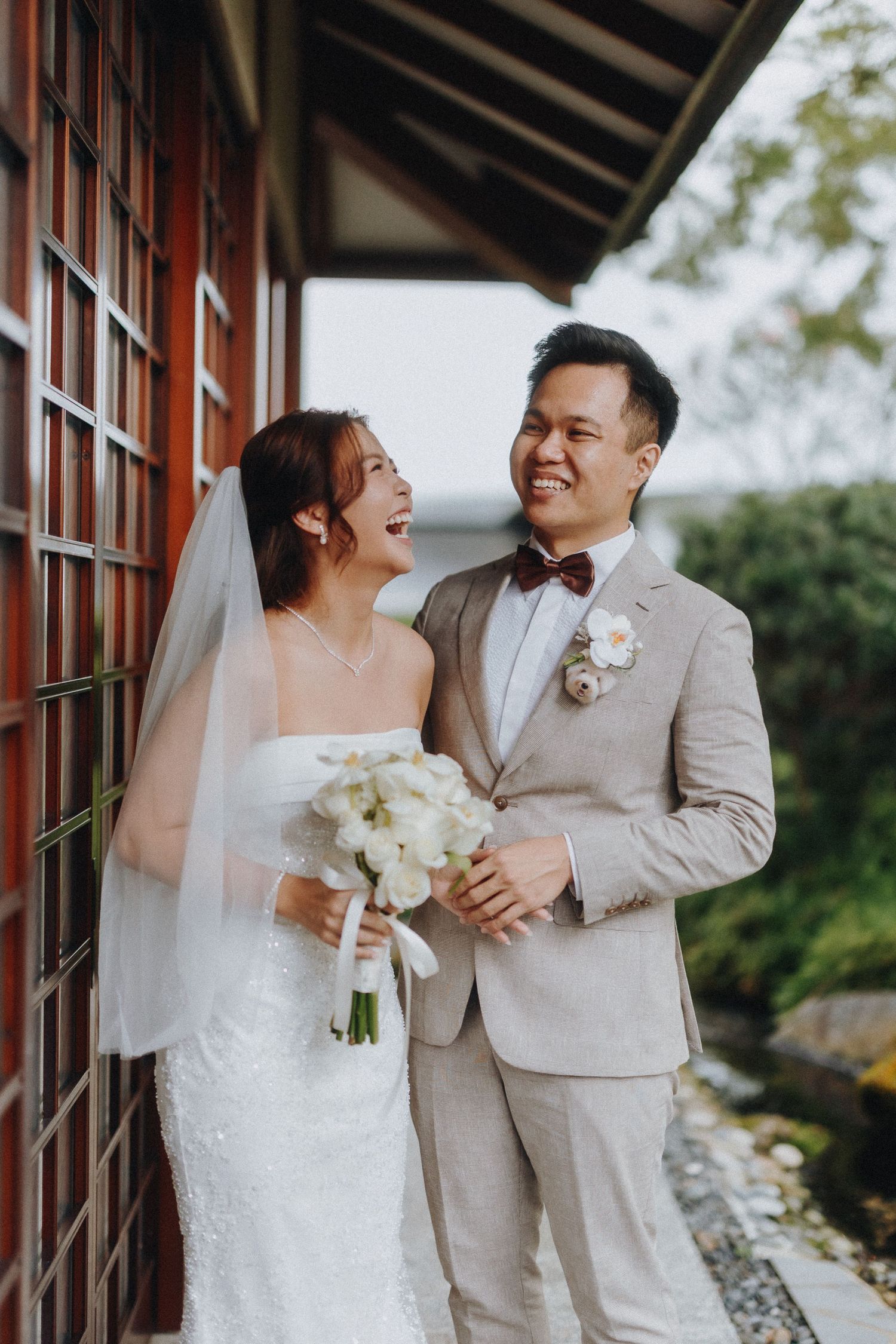 A wedding couple in matching light beige and white attire share a joyful candid moment by a rustic wooden wall.