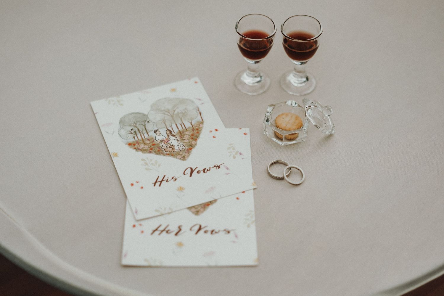 Wedding rings and glasses of red wine with wedding stationery featuring rose gold text on a white table surface.