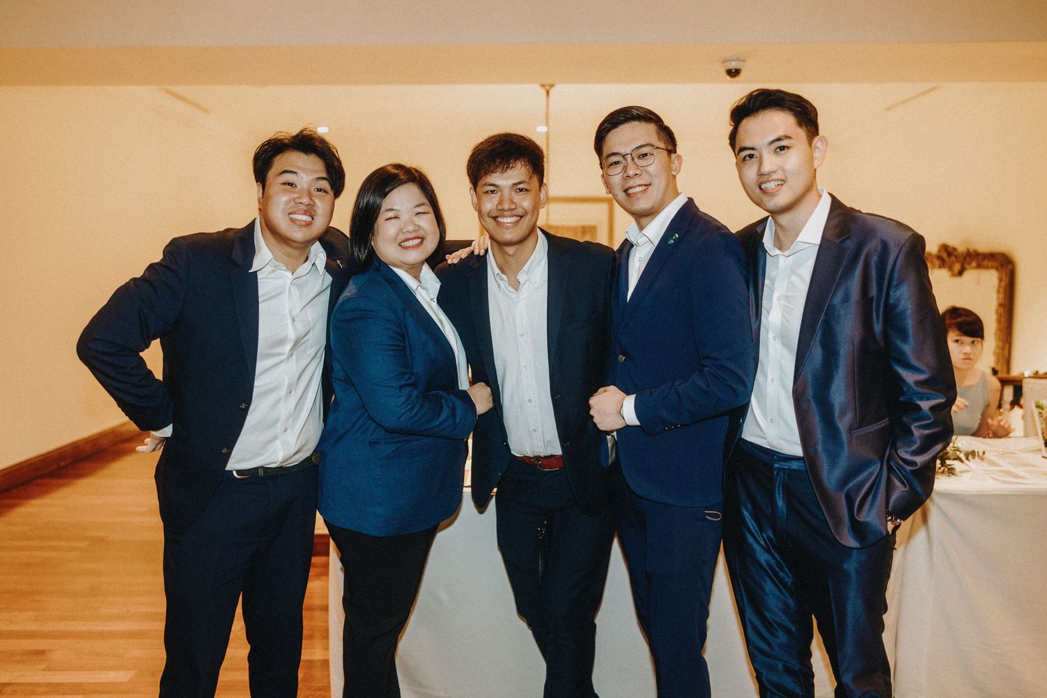 Group photo of wedding party members in navy suits and formal attire in an indoor venue.