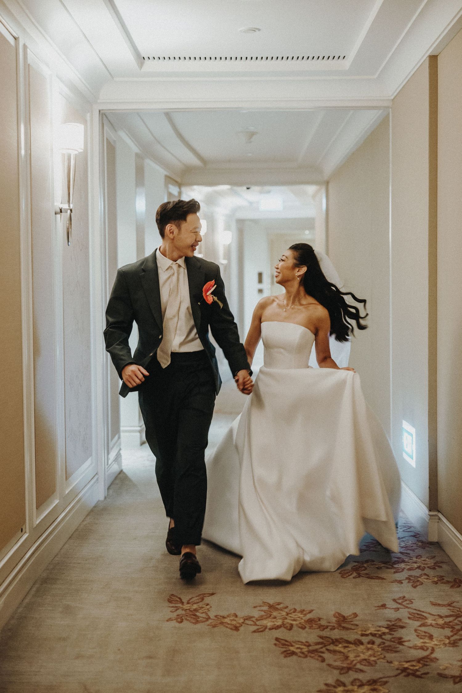A couple in wedding attire playfully running down an elegant hotel hallway with white walls and floral carpeting.