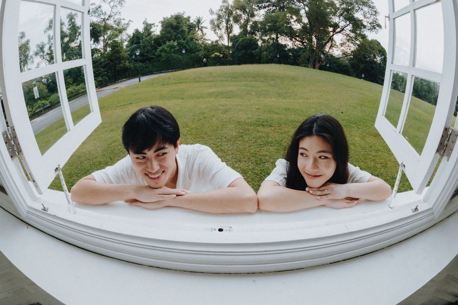 A creative fisheye view through a circular window frame showing two young people leaning on the windowsill outdoors.