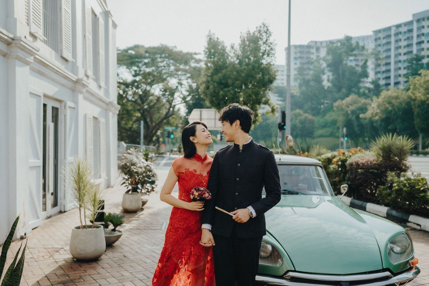 A couple in formal wear poses next to a vintage mint green car on a sunny street.