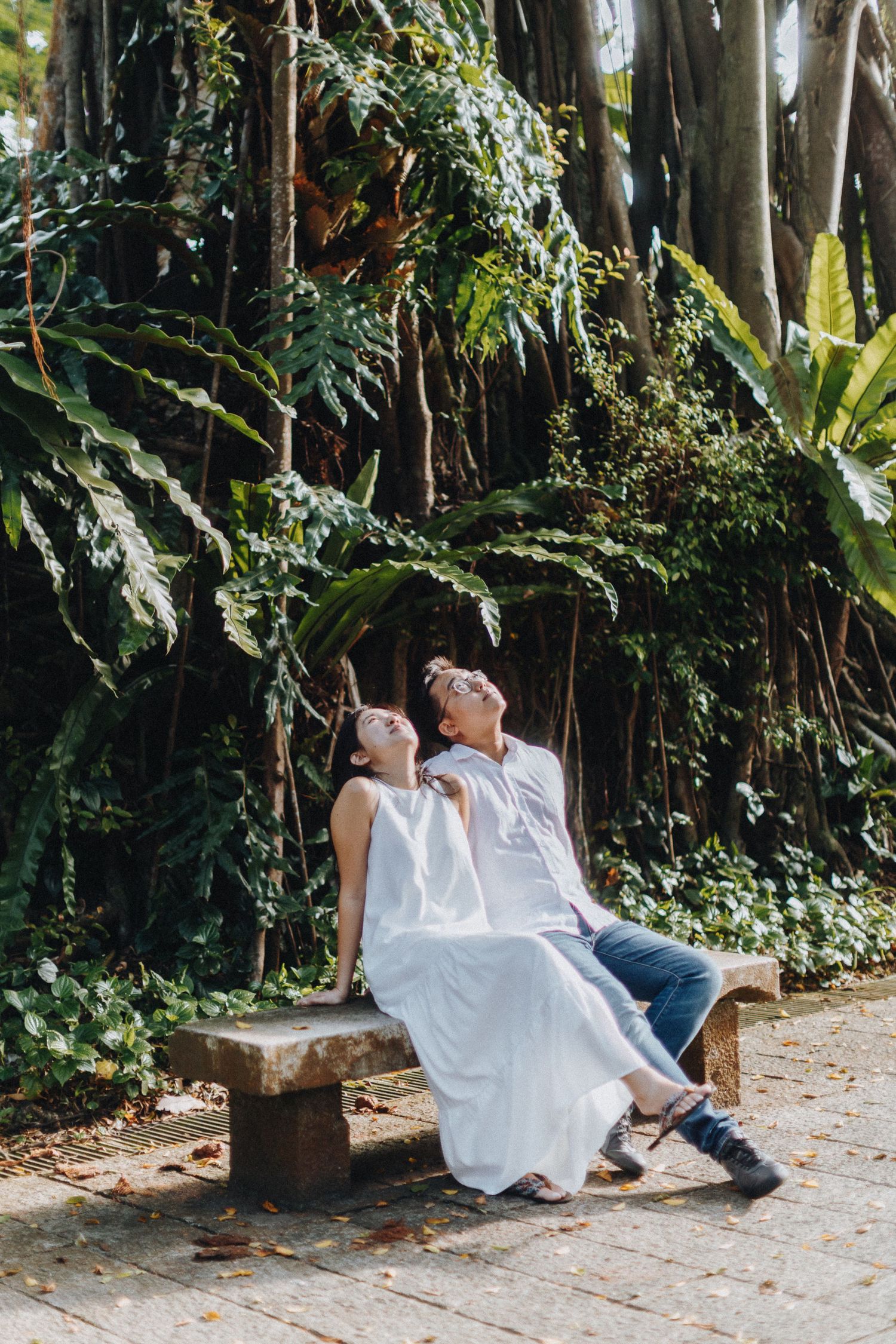 A couple sits together on a bench surrounded by tropical plants and trees.