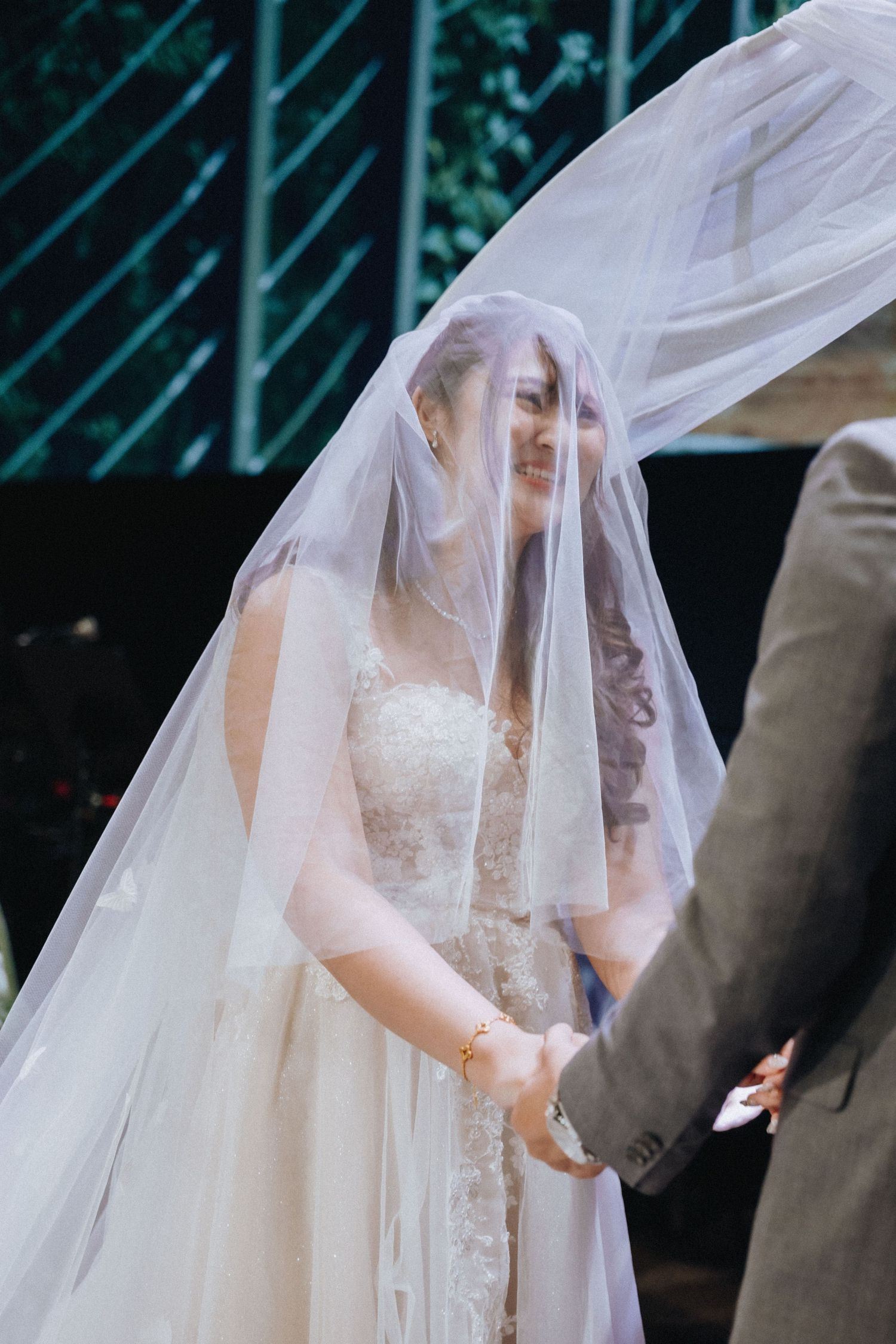 A bride in a flowing white wedding dress and long veil stands outdoors with greenery in the background.