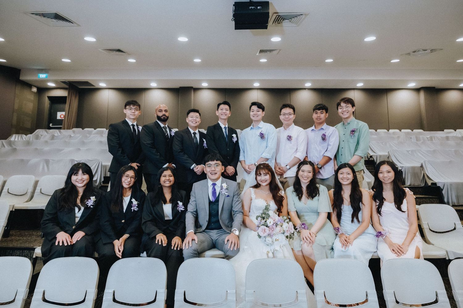 Wedding party members in gray suits and white dresses seated in rows for a formal group photo.