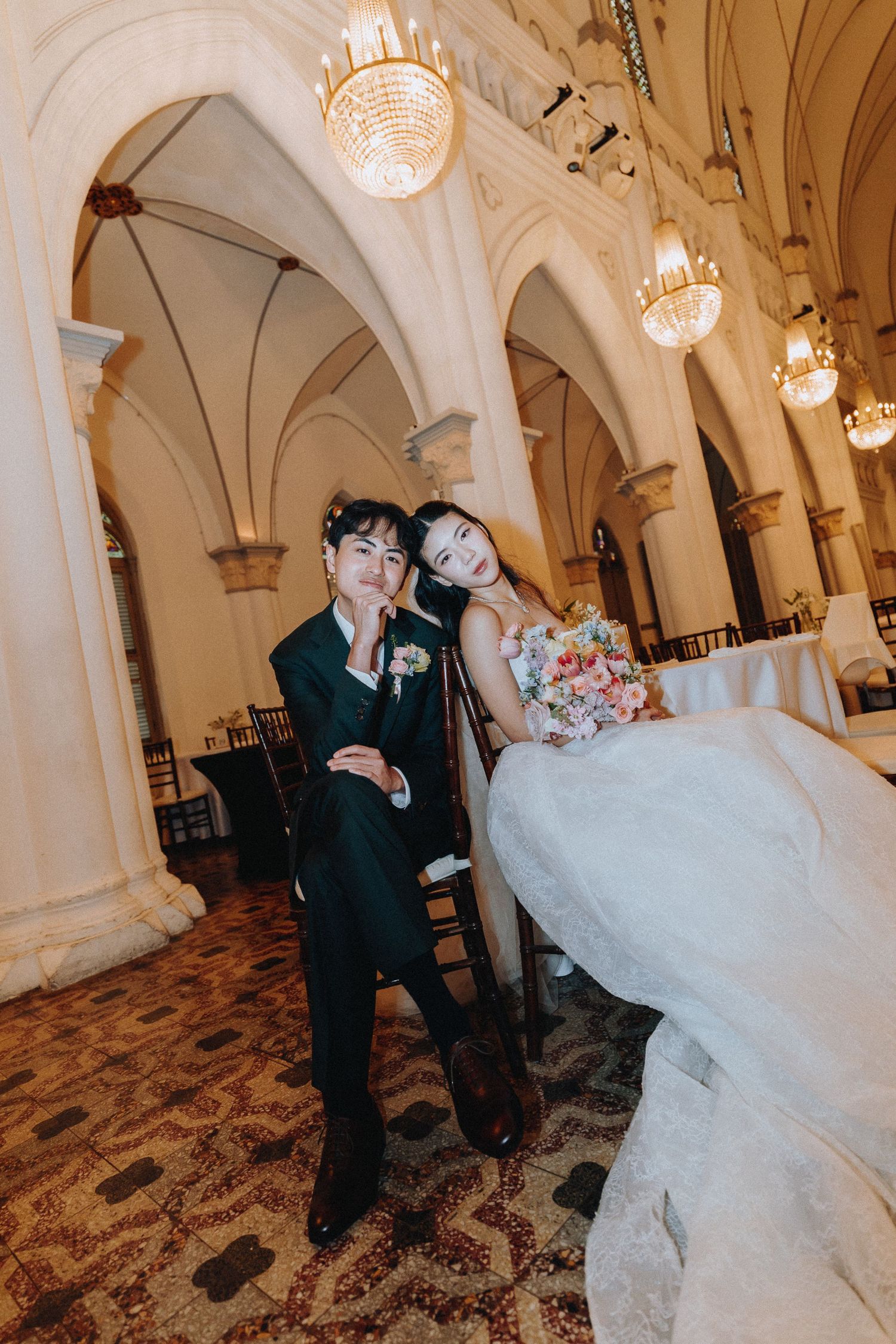 Elegant wedding couple posing in ornate venue with arched ceilings and crystal chandeliers, bride holding colorful bouquet.