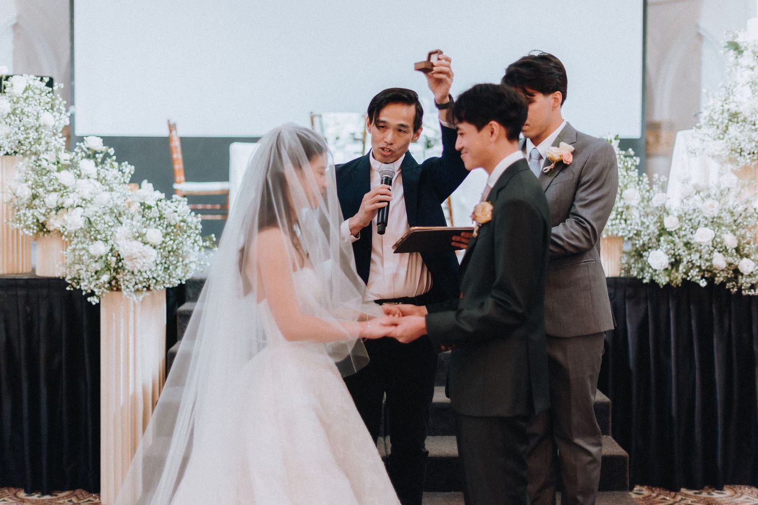 Couple exchanging wedding rings during a ceremony officiated by a minister, with floral arrangements in the background.