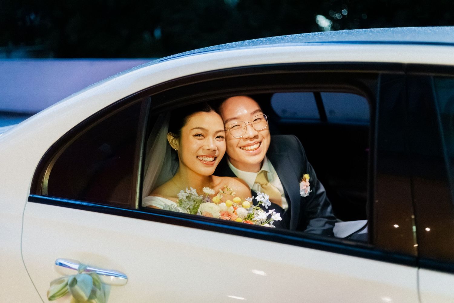 Smiling bride and groom peering through car window on wedding day, bride holding bouquet.