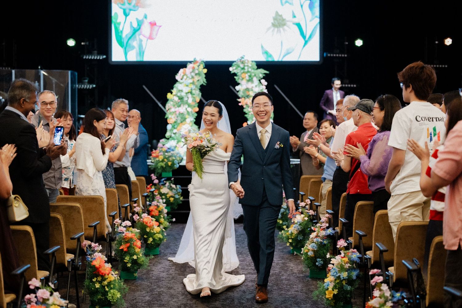 Newlyweds walk down the aisle as guests applaud at a vibrant floral-decorated wedding reception.
