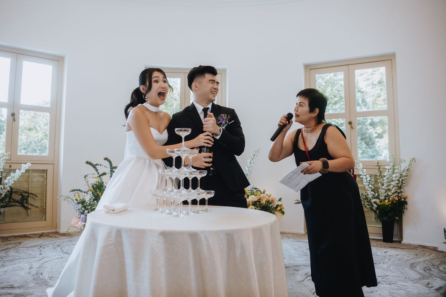 Bride and groom laughing beside a champagne tower while a woman gives a speech.