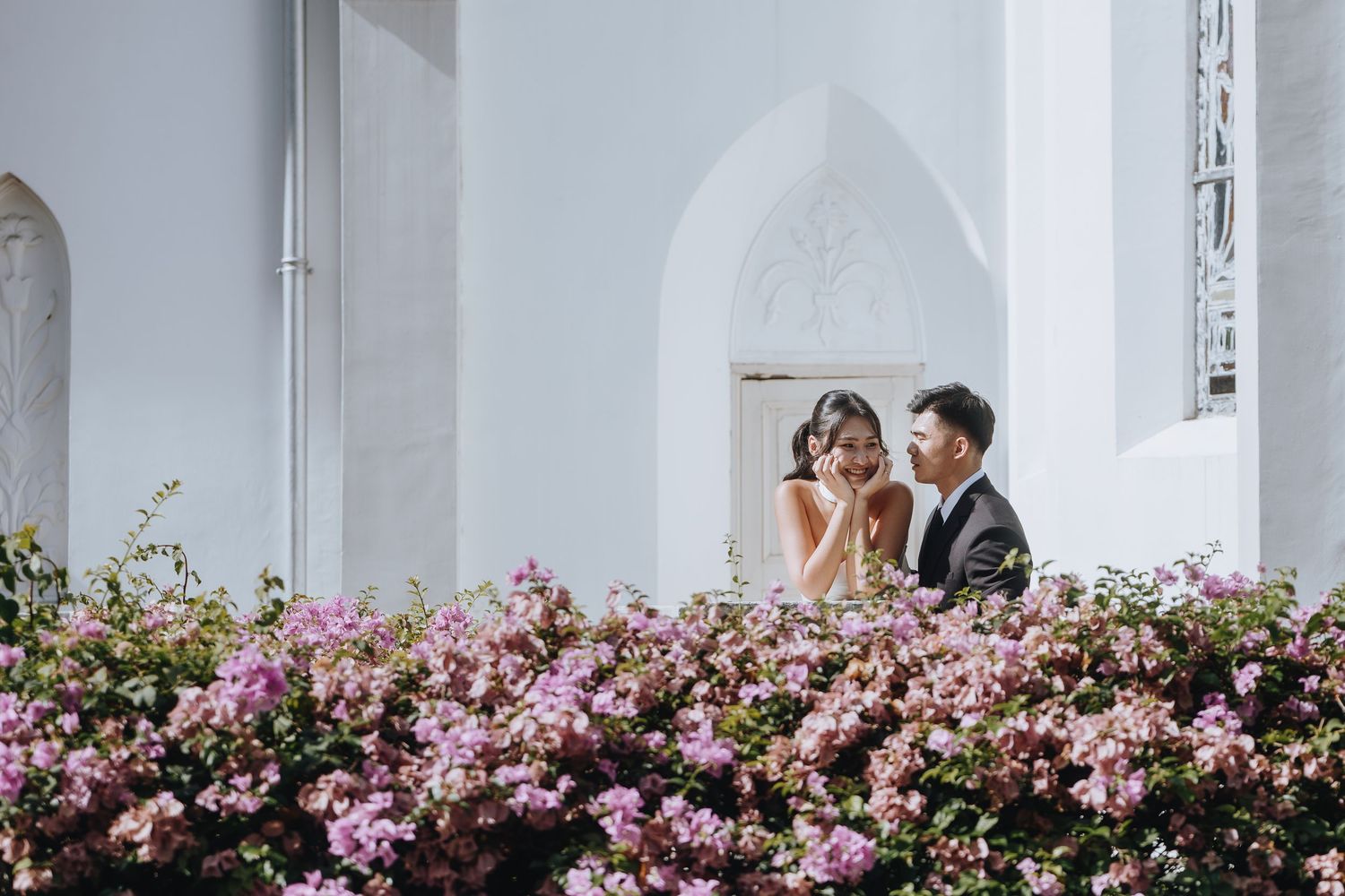 Bride and groom share a romantic moment on a balcony surrounded by blooming pink bougainvillea flowers.