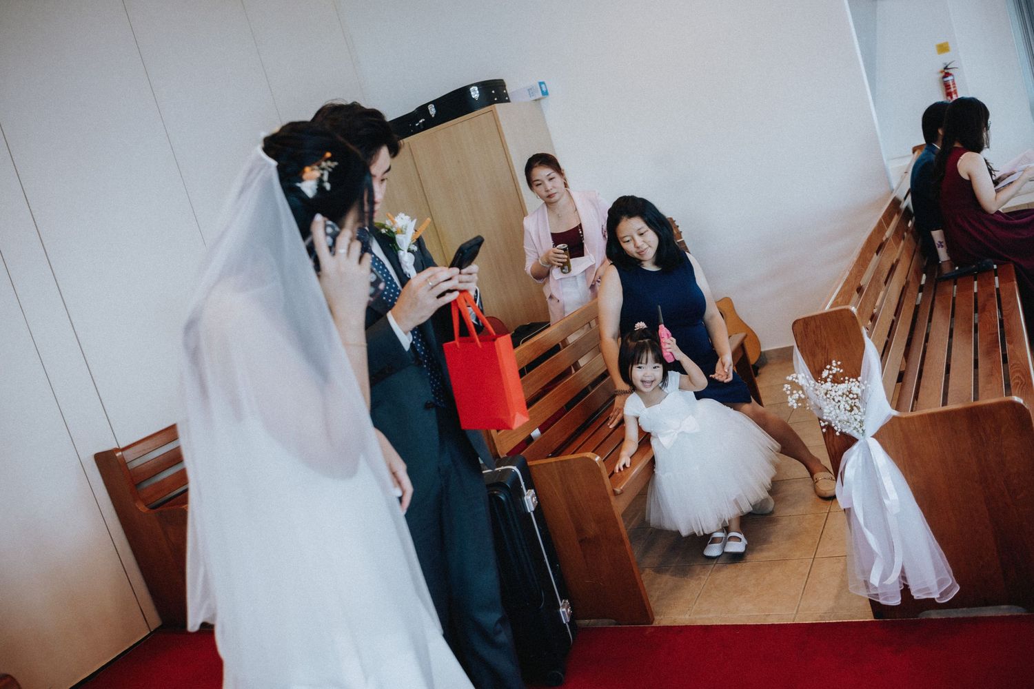 Bride in white gown with wedding guests in church pews, flower girl in white dress during wedding ceremony.