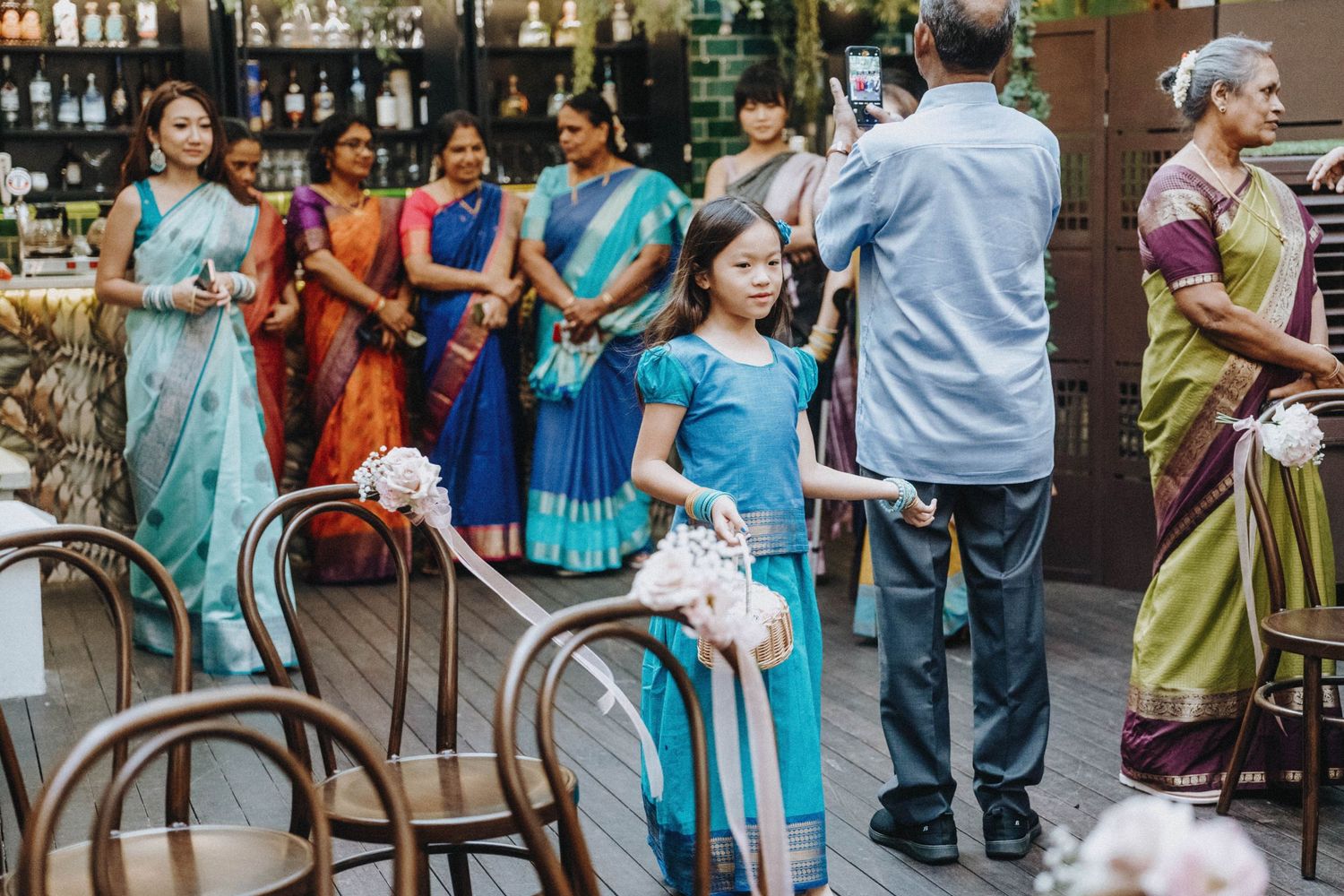 Women in colorful saris stand at an Indian wedding ceremony outdoors, with a young girl holding white flowers.