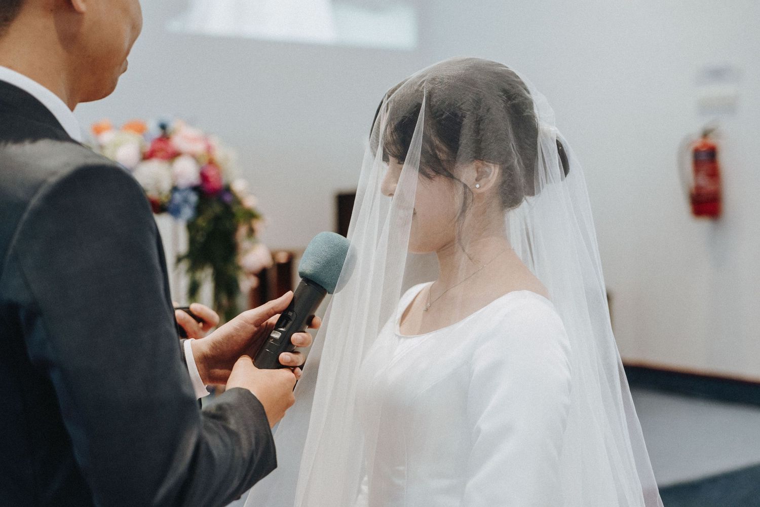 Bride in white gown and veil holding microphone during intimate wedding ceremony vows.