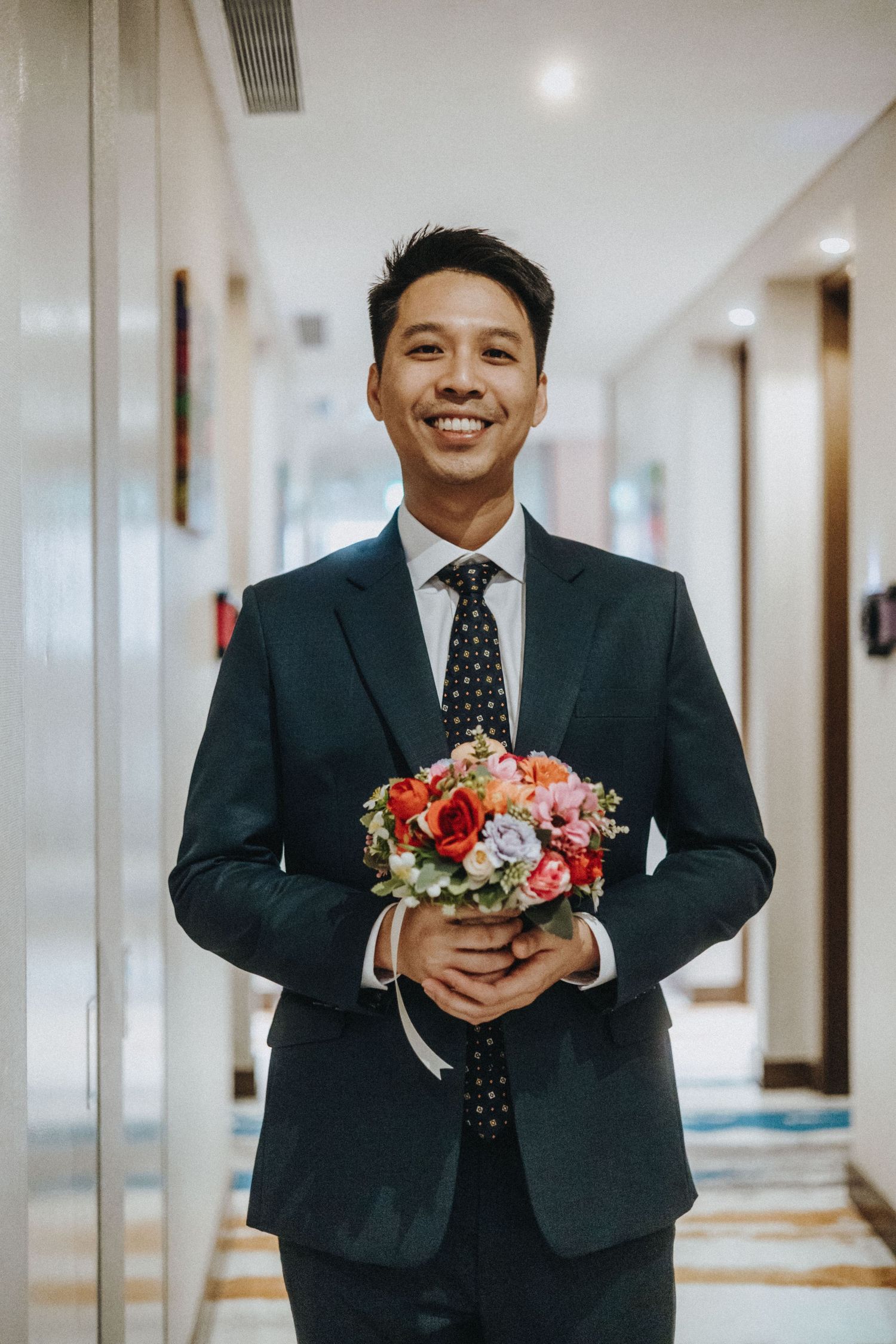 Smiling groom in dark suit holding colorful bridal bouquet in hotel corridor.