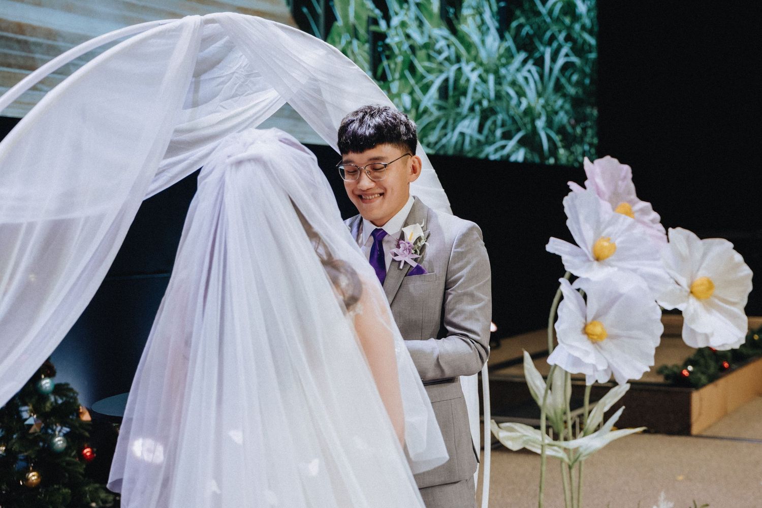 Wedding ceremony setup with flowing white veil against lush green foliage backdrop and white floral arrangements.