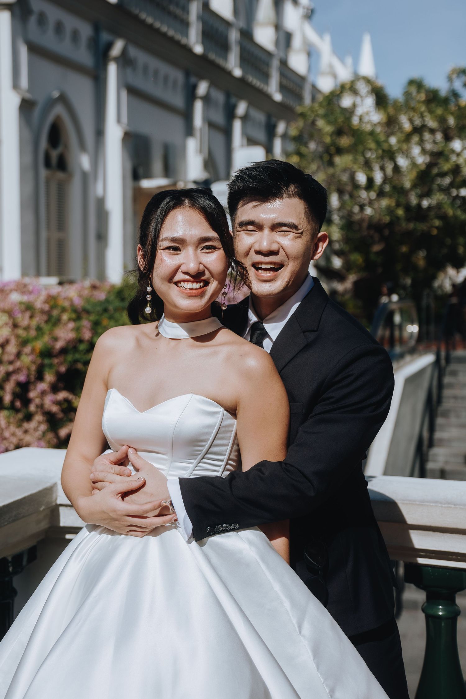 Smiling bride and groom embracing outdoors in wedding attire near a historic building.