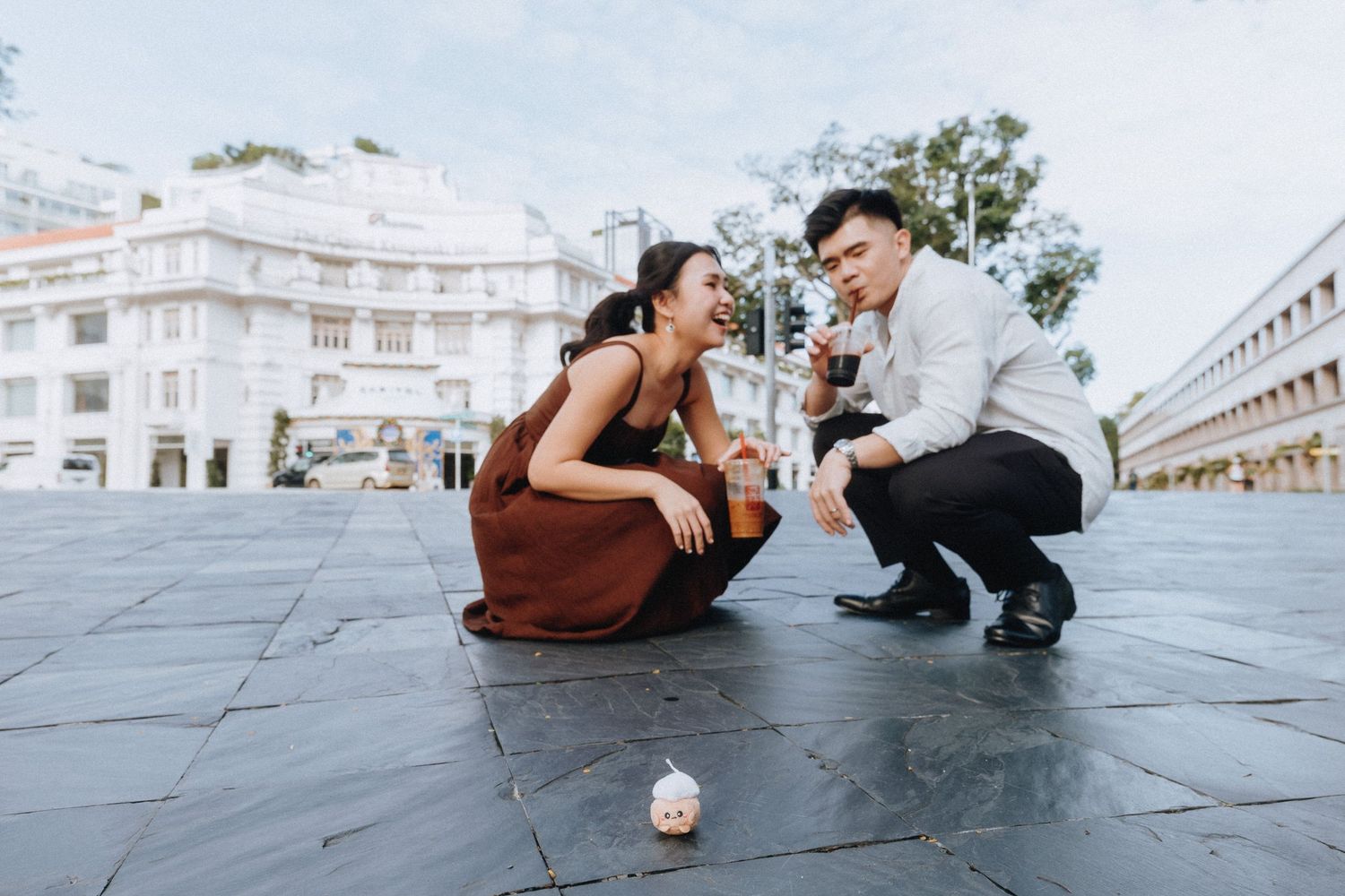 A man and woman crouch on a city plaza examining a small crumpled object on the ground, dressed in elegant attire.