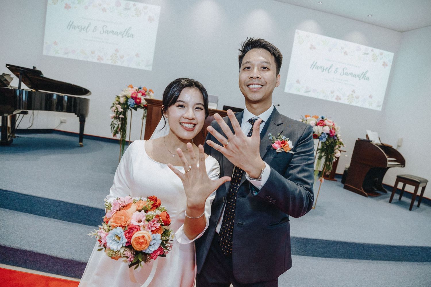 A smiling bride and groom show off their wedding rings inside a decorated church chapel.