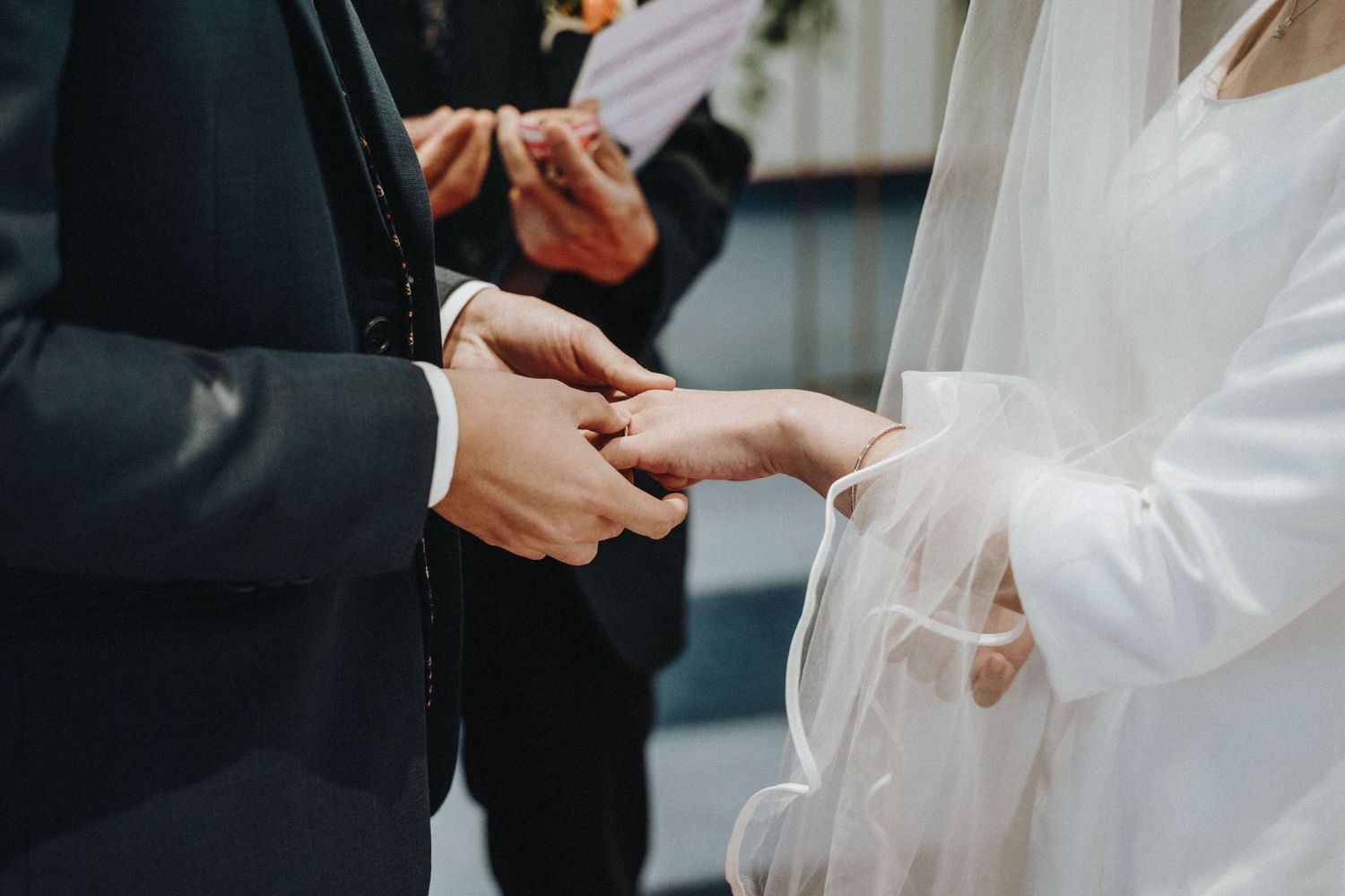 Groom places wedding ring on brides finger during an outdoor wedding ceremony with officiant in background.