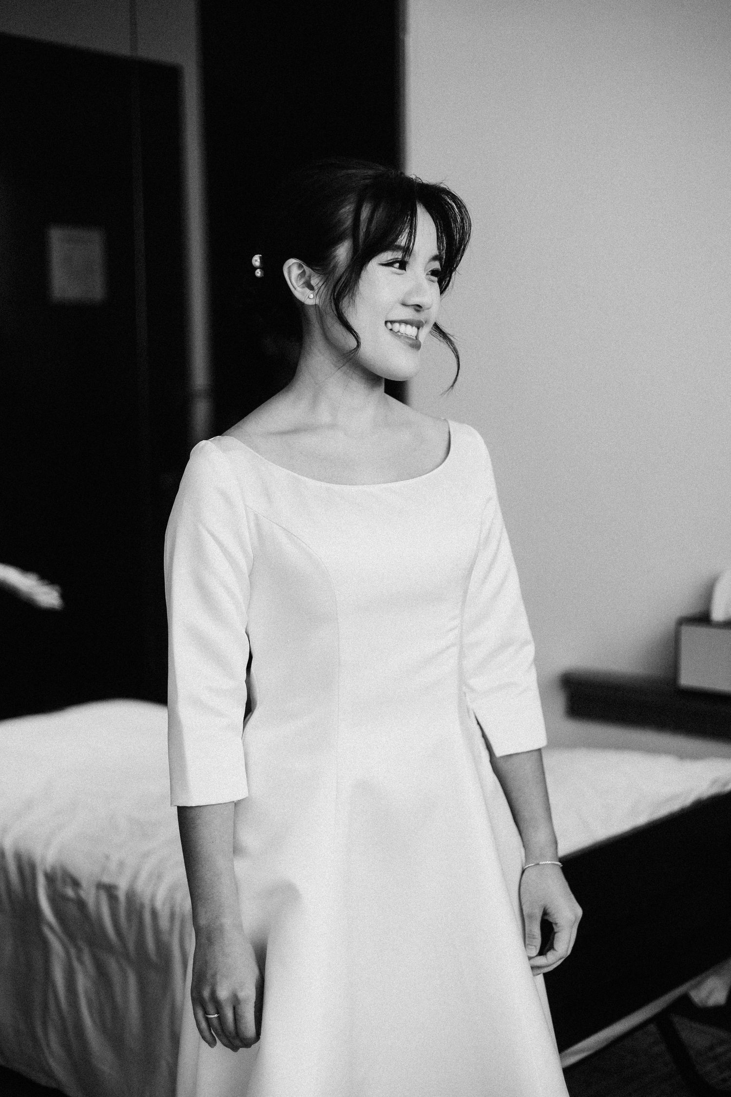 Smiling bride in elegant white dress with three-quarter sleeves poses in a hotel room in black and white photo.