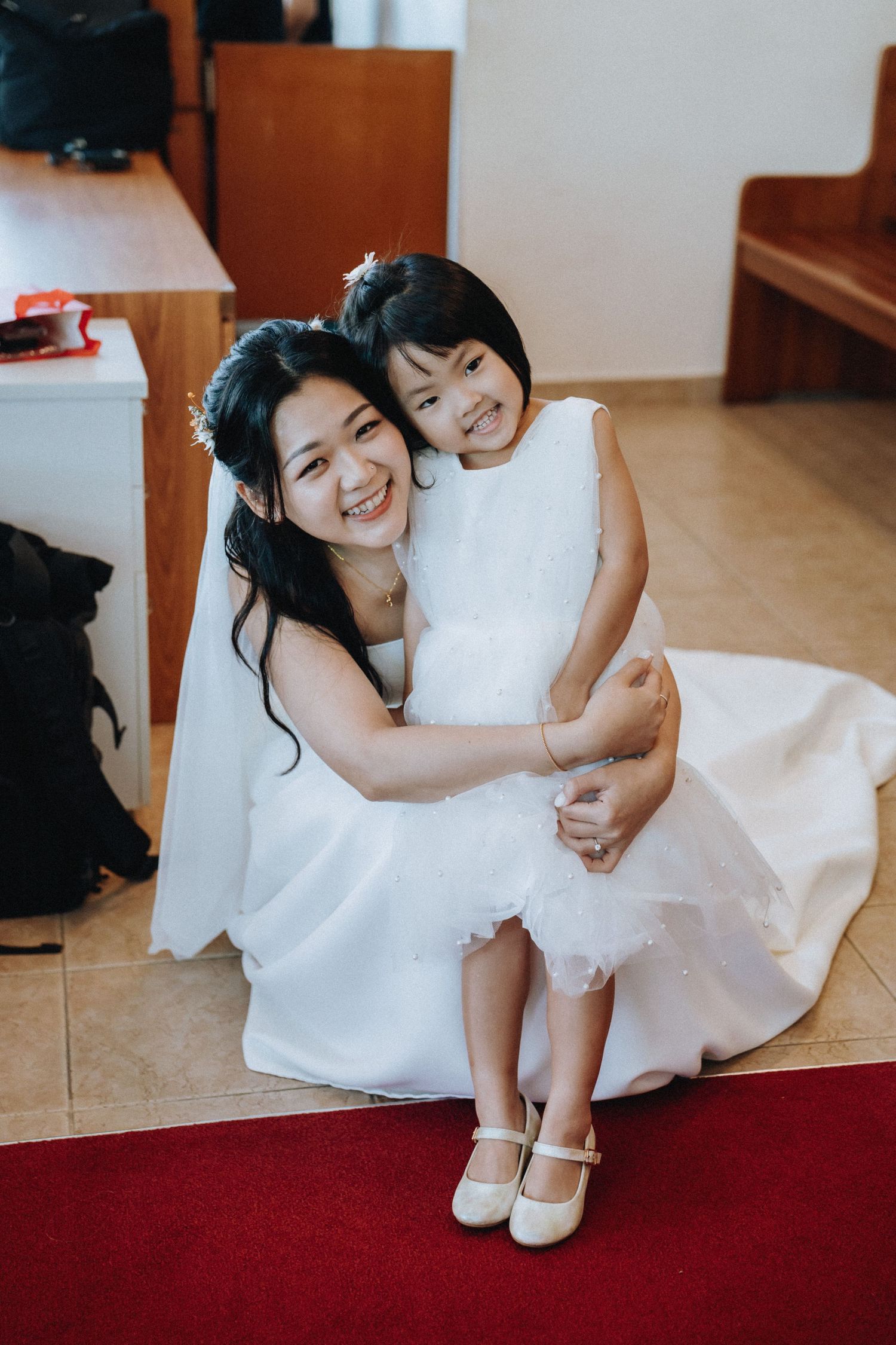 Smiling bride in white gown hugs a young flower girl in a white dress on a red carpet in a church.