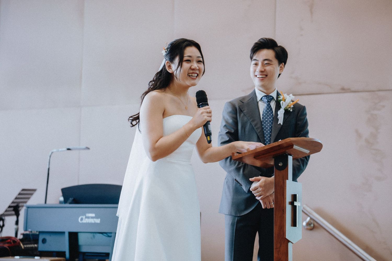 Smiling bride speaks into microphone at wedding ceremony while groom stands beside her at the podium.